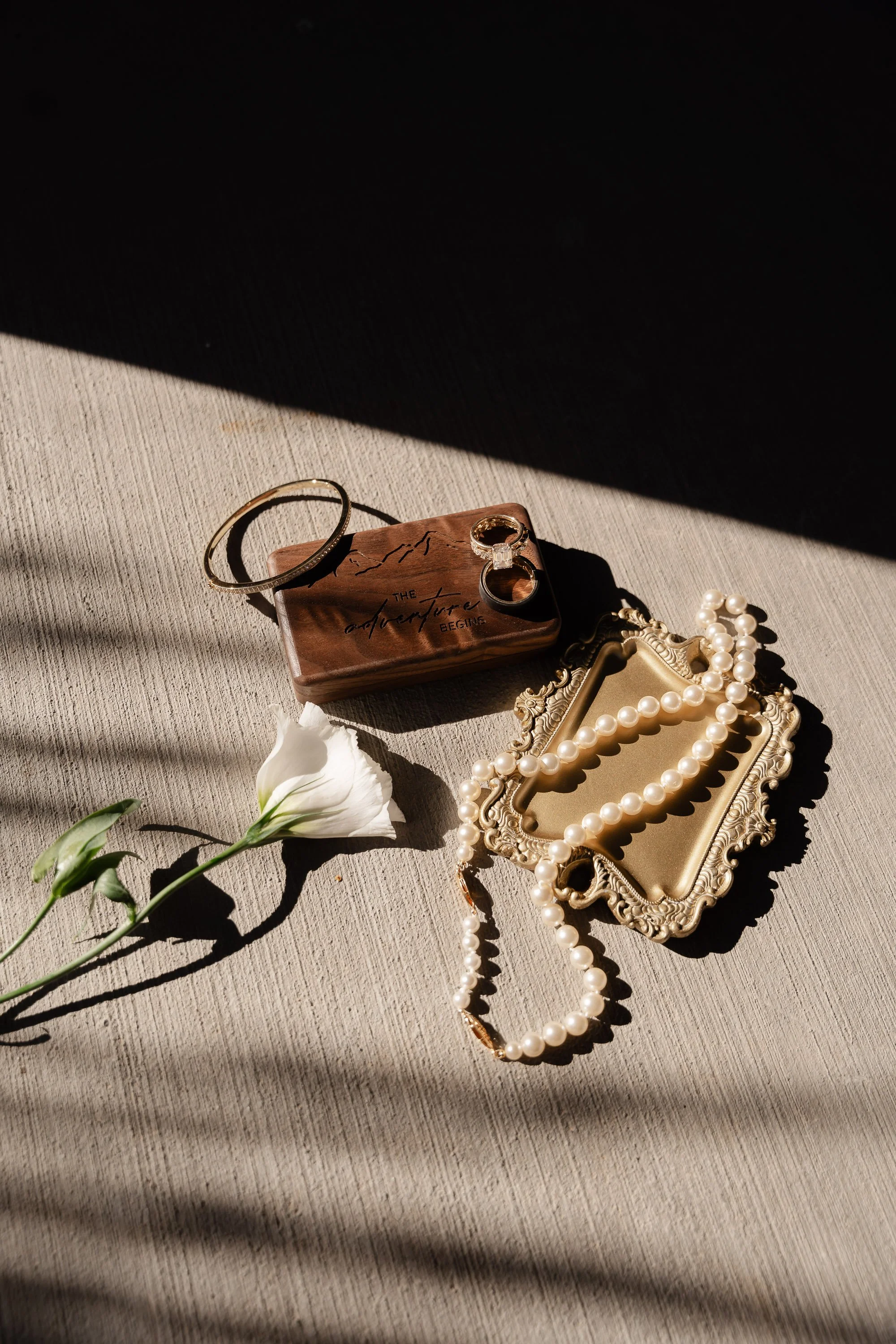 Jewelry and accessories arranged on a textured fabric surface, including a pearl necklace, a pearl bracelet, a gold ring, a gold bracelet, a decorative mirror, a flower, and a small jewelry box.