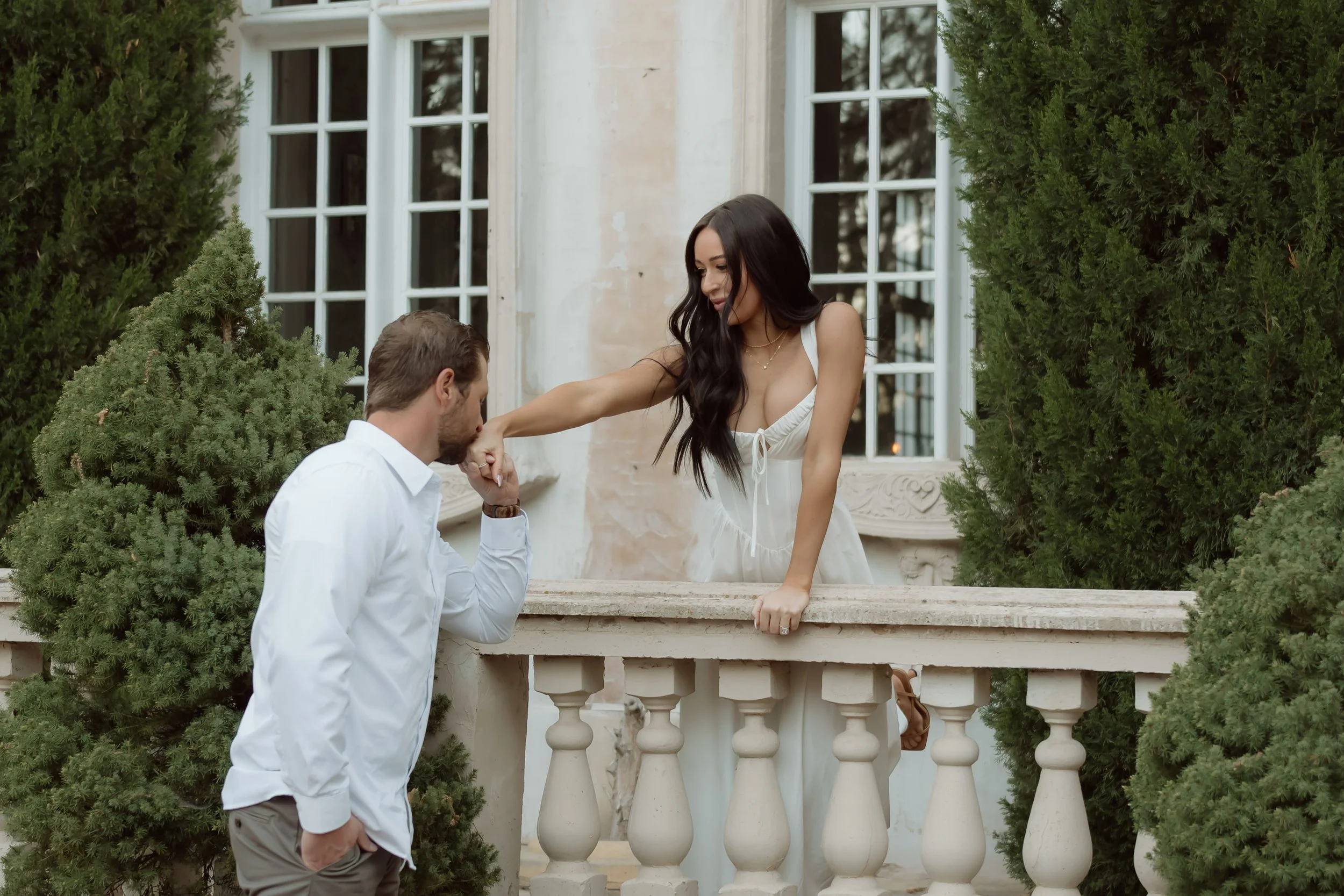 A man kneeling and holding a woman's hand on a balcony railing, with the woman leaning on the railing, in front of a large window and surrounded by greenery.