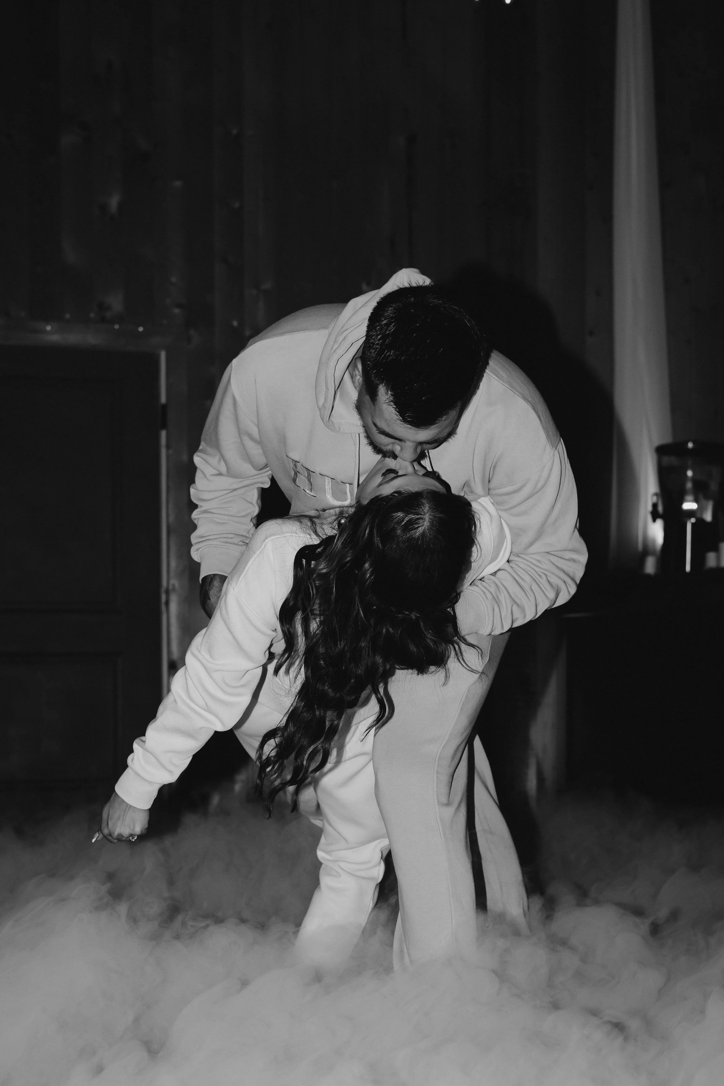 A man and woman kissing while dancing on a wooden floor, surrounded by fog with a dark wooden wall in the background.