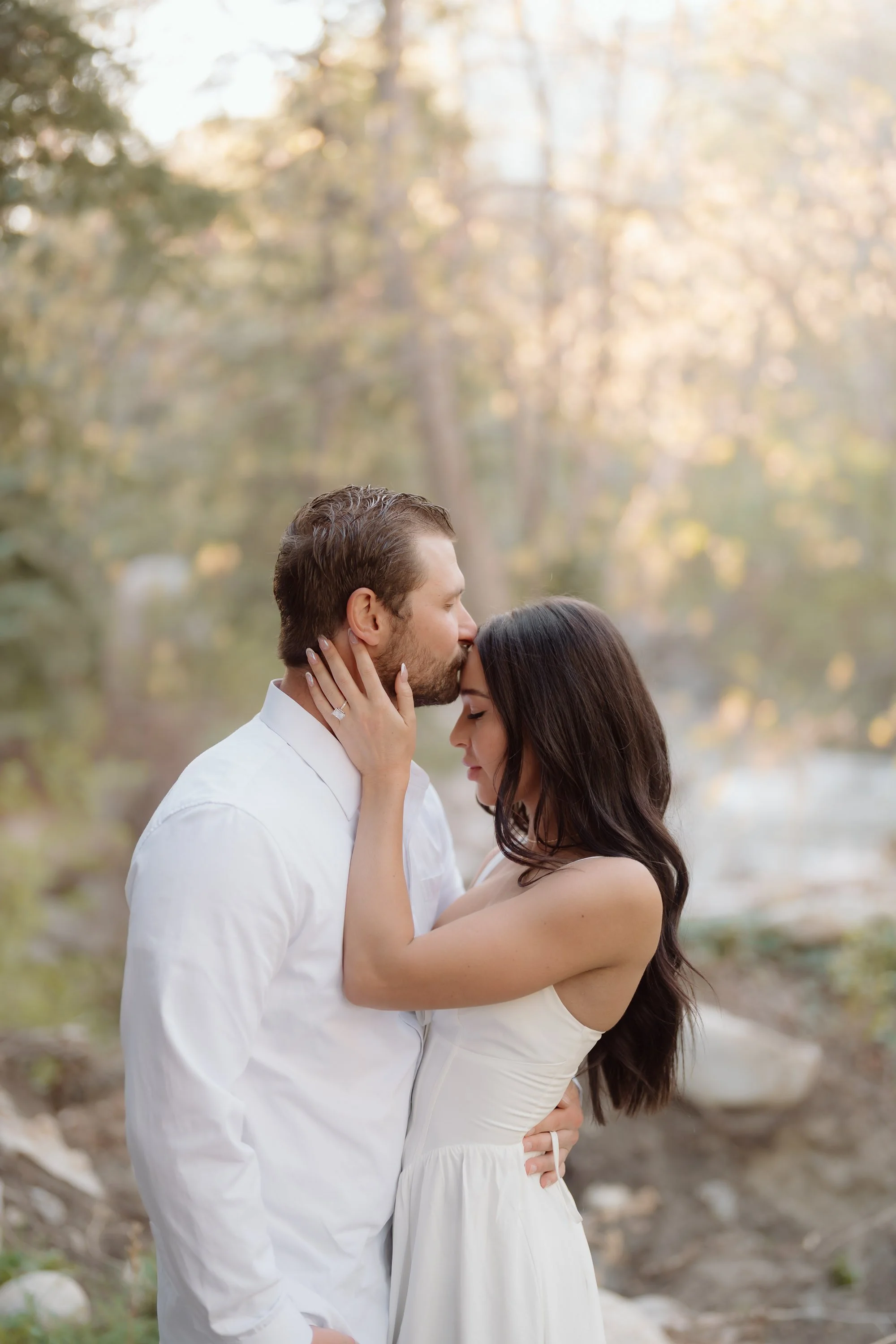 A couple embraces and kisses in a natural setting with trees and soft light.