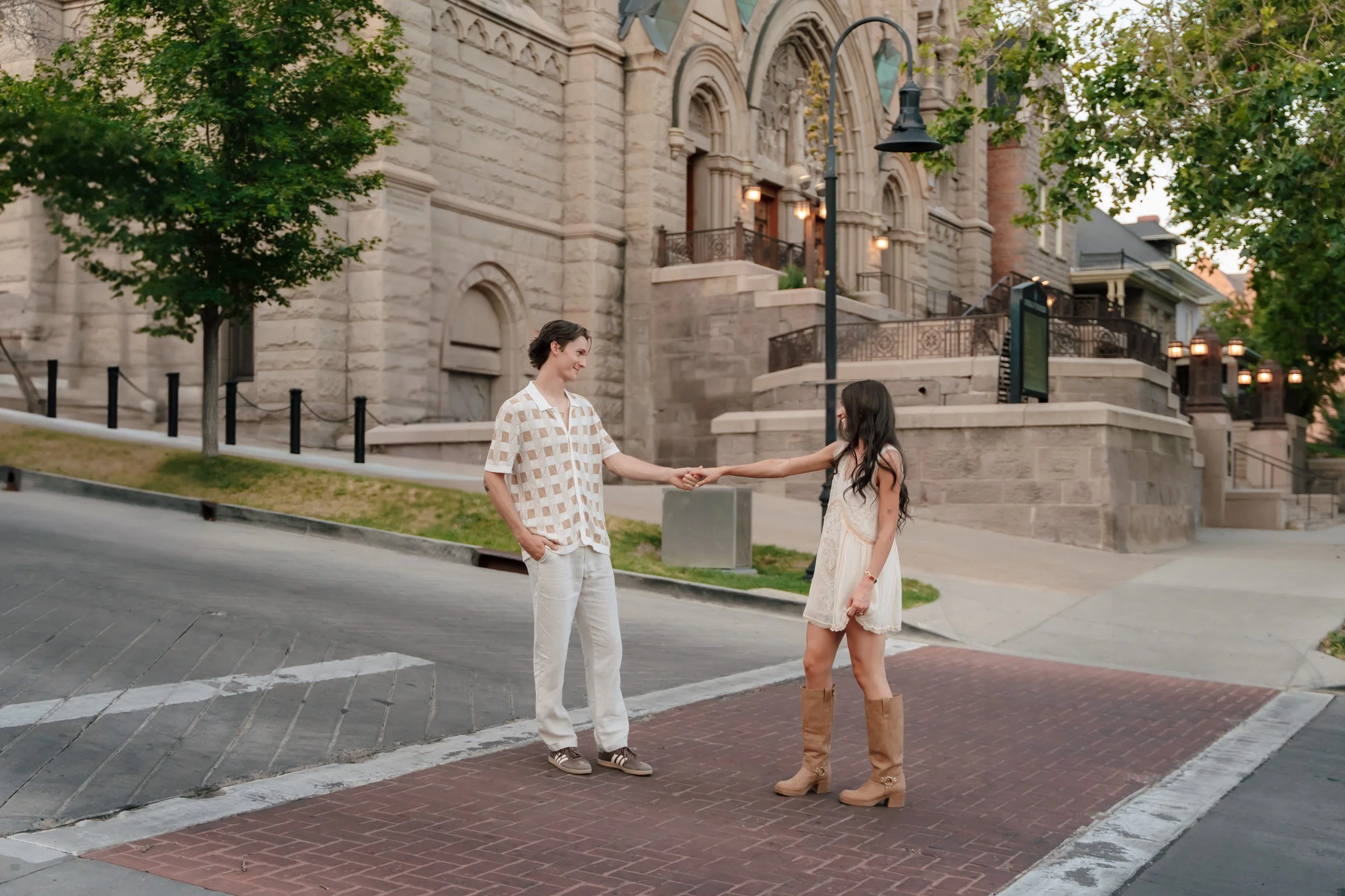 A man and a woman holding hands on a city sidewalk in front of a historic stone building with arched windows and stairs. Trees and street lamps are visible.