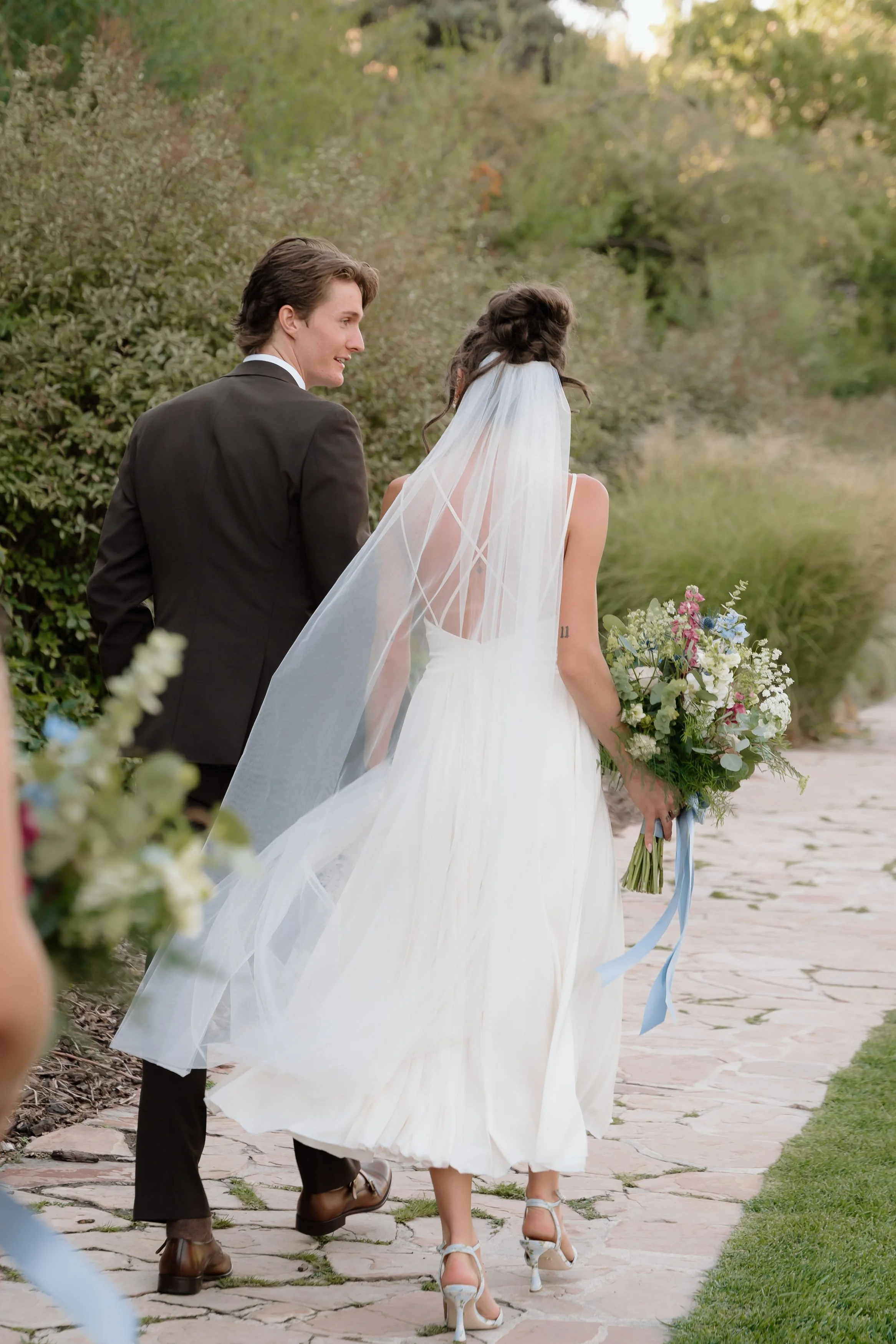 A bride in a white wedding dress and a groom in a black suit walking outdoors on a stone path, holding a bouquet of flowers, surrounded by greenery.