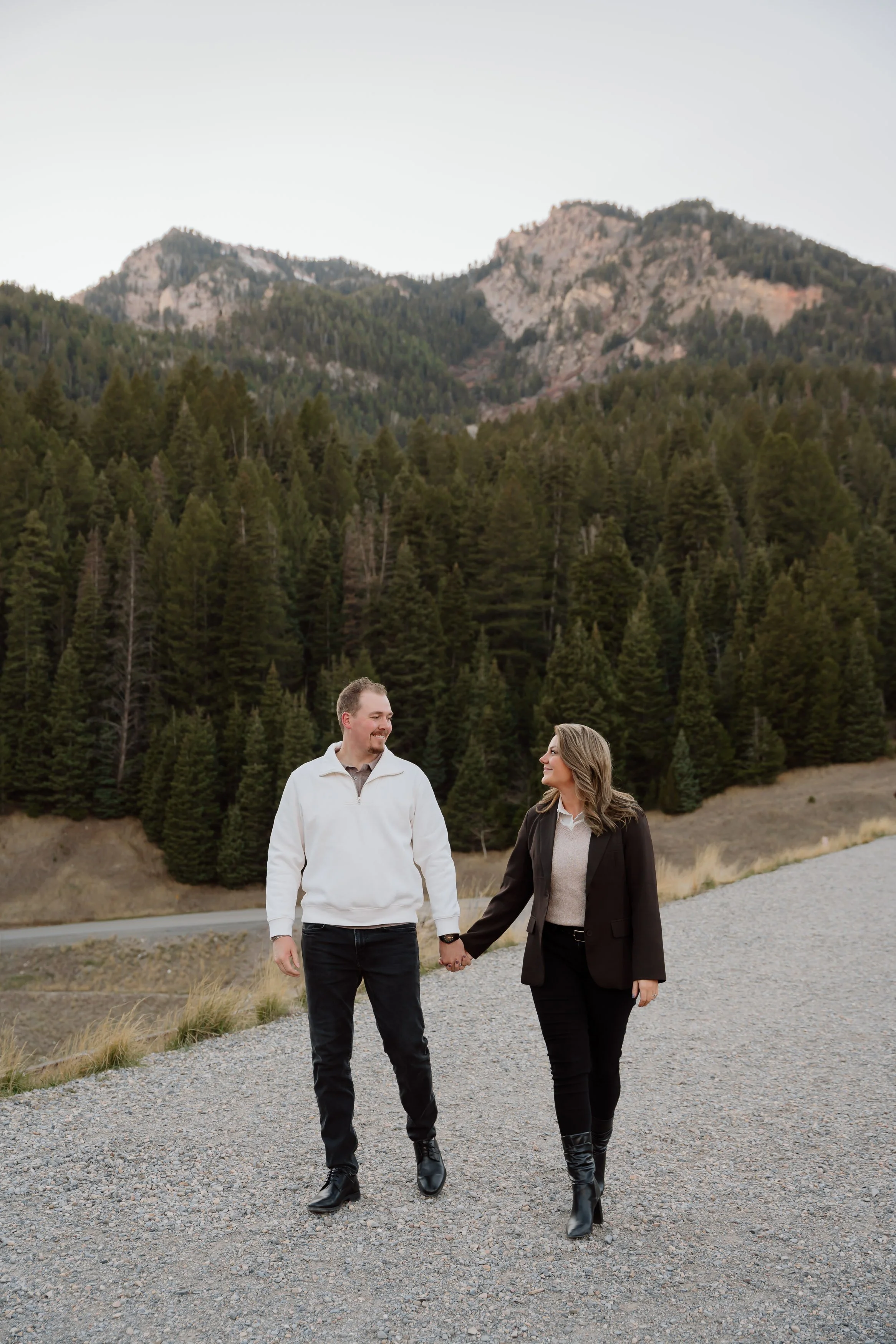 A man and a woman walking hand in hand on a gravel road in a mountainous forested area, with mountains in the background.