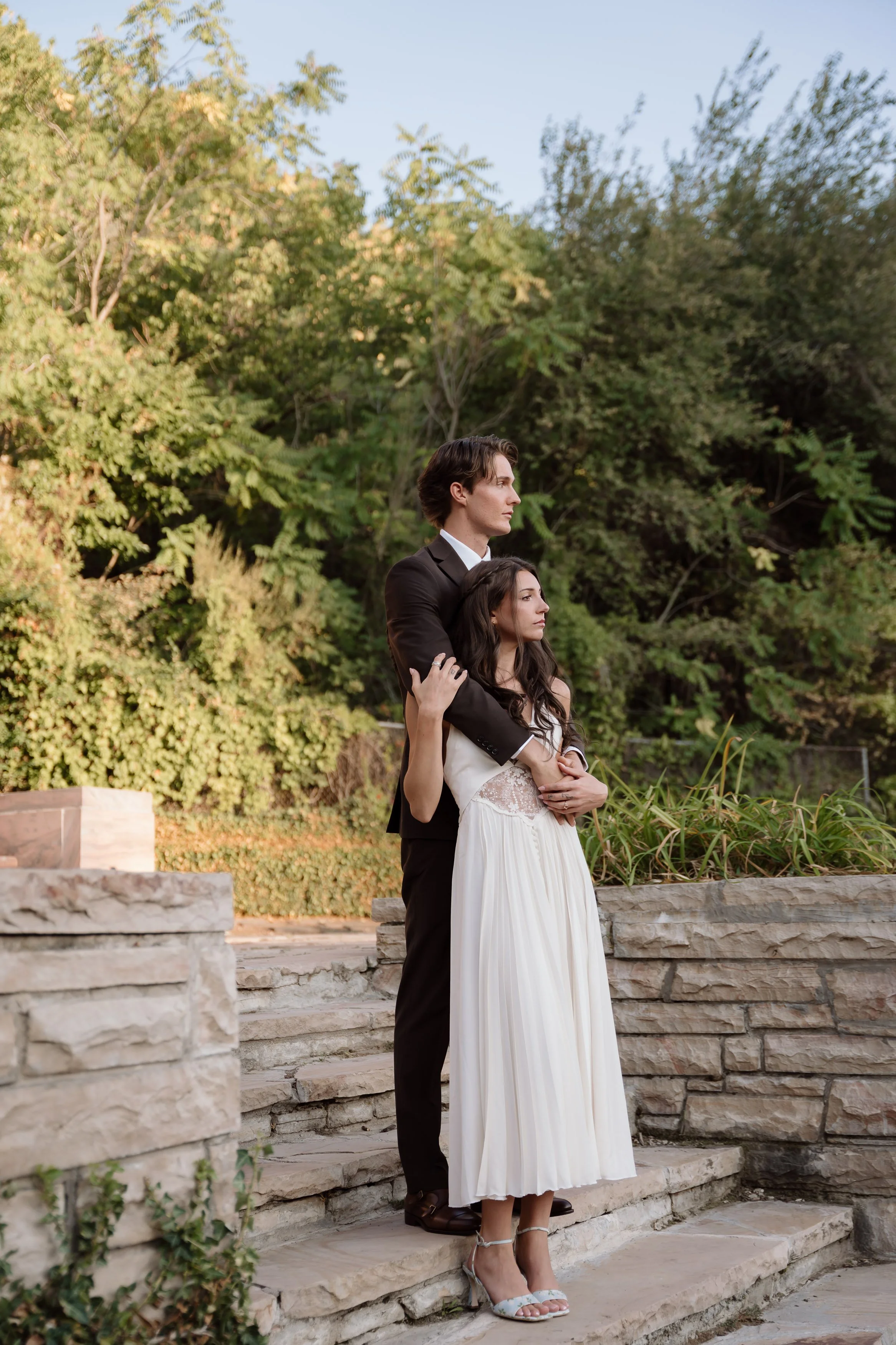 A couple standing on stone steps outdoors surrounded by greenery, with the man dressed in a suit and the woman in a white dress, embracing each other.