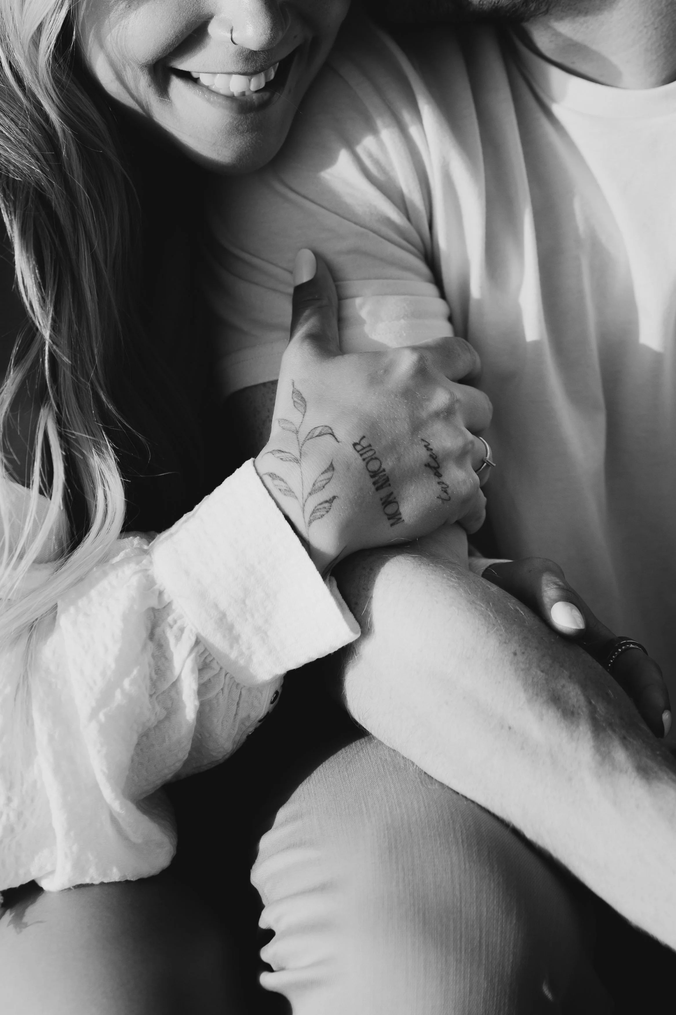 Close-up of a smiling woman's face with a nose ring, holding a man's penis, with tattoos on her hand that say 'MOM MOM', and the woman is wearing a white shirt, in black and white.