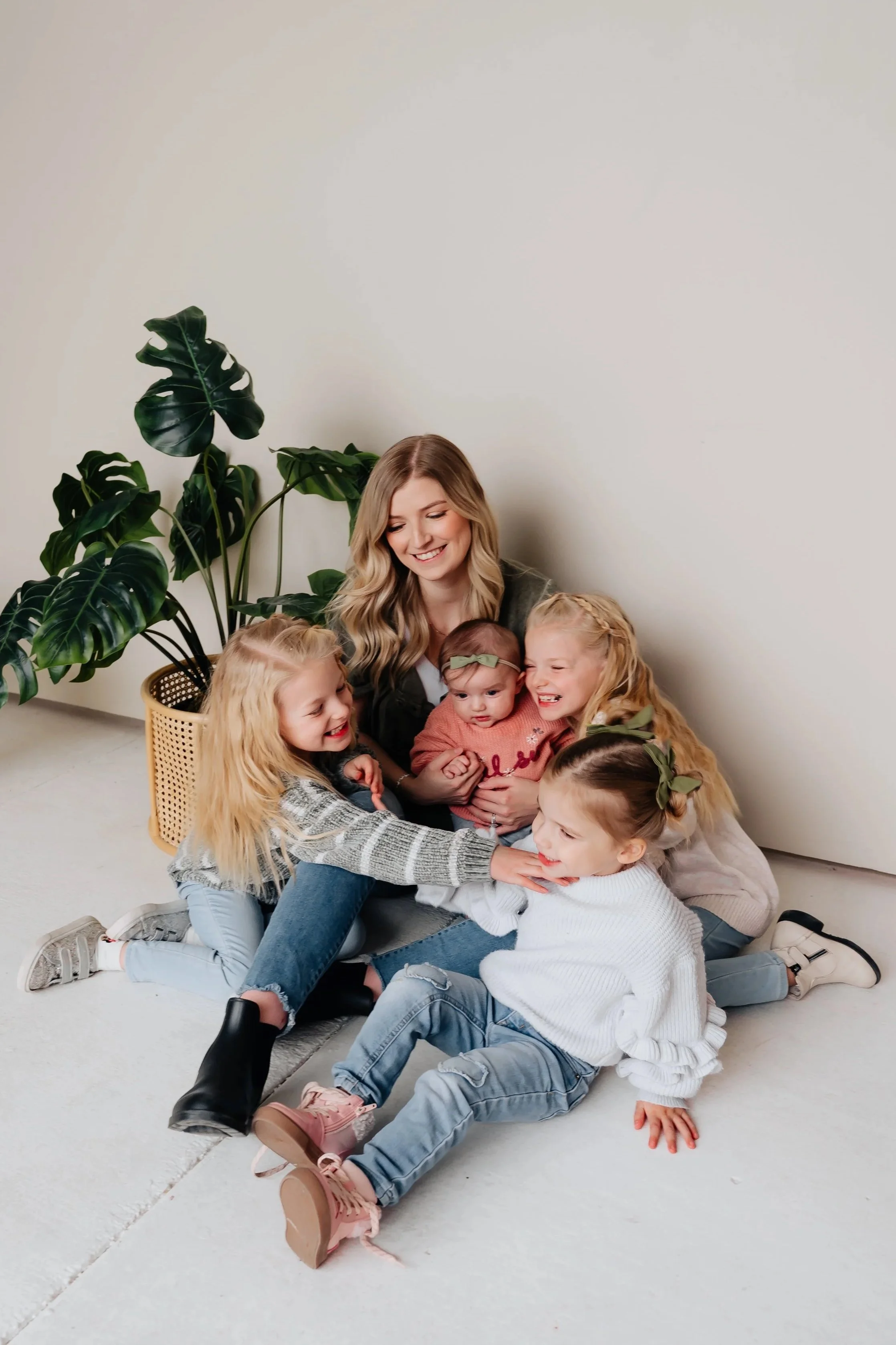 A woman sitting on the floor surrounded by five young girls, smiling and playing together in a bright room with a large green plant near the wall.