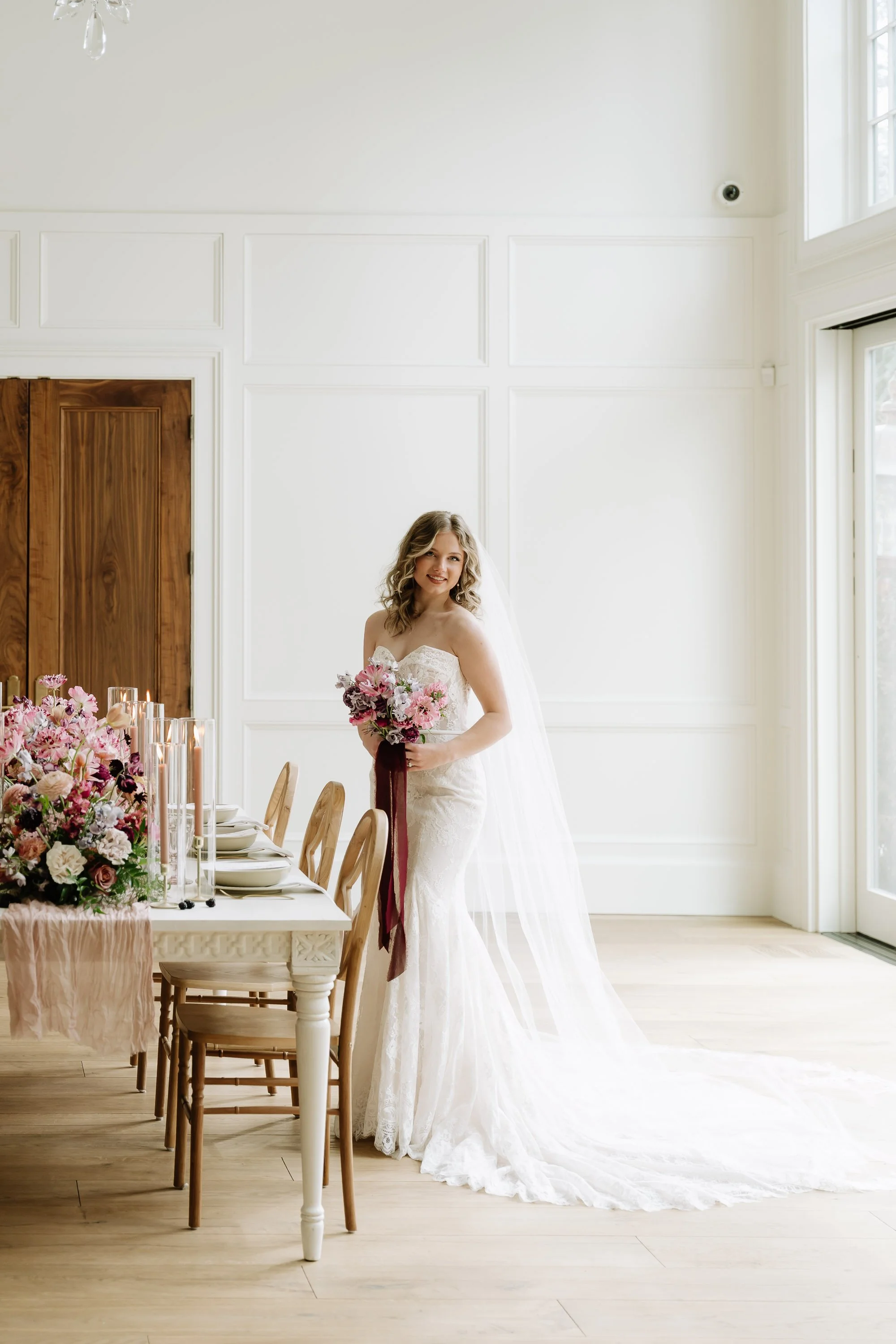 A bride in a white lace wedding dress holding a bouquet of pink and purple flowers, standing beside a dining table decorated with pink flowers and candles in glass holders, in a bright, white room with large windows.