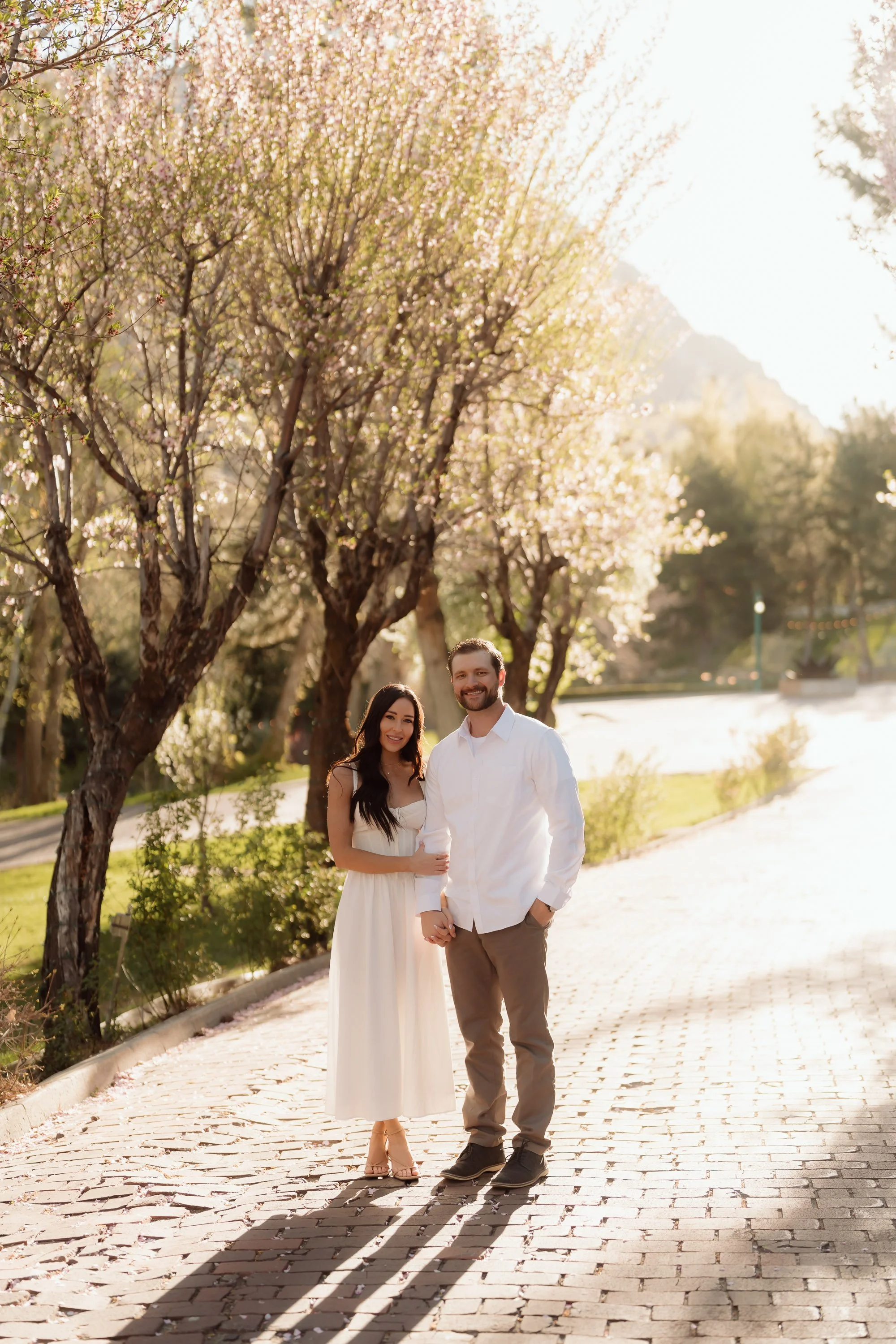 A young couple stands on a cobblestone path under blooming trees in sunlight. The woman has long dark hair, is wearing a white dress, and the man has a beard, is wearing a white shirt and beige pants. They are holding hands and smiling at the camera.