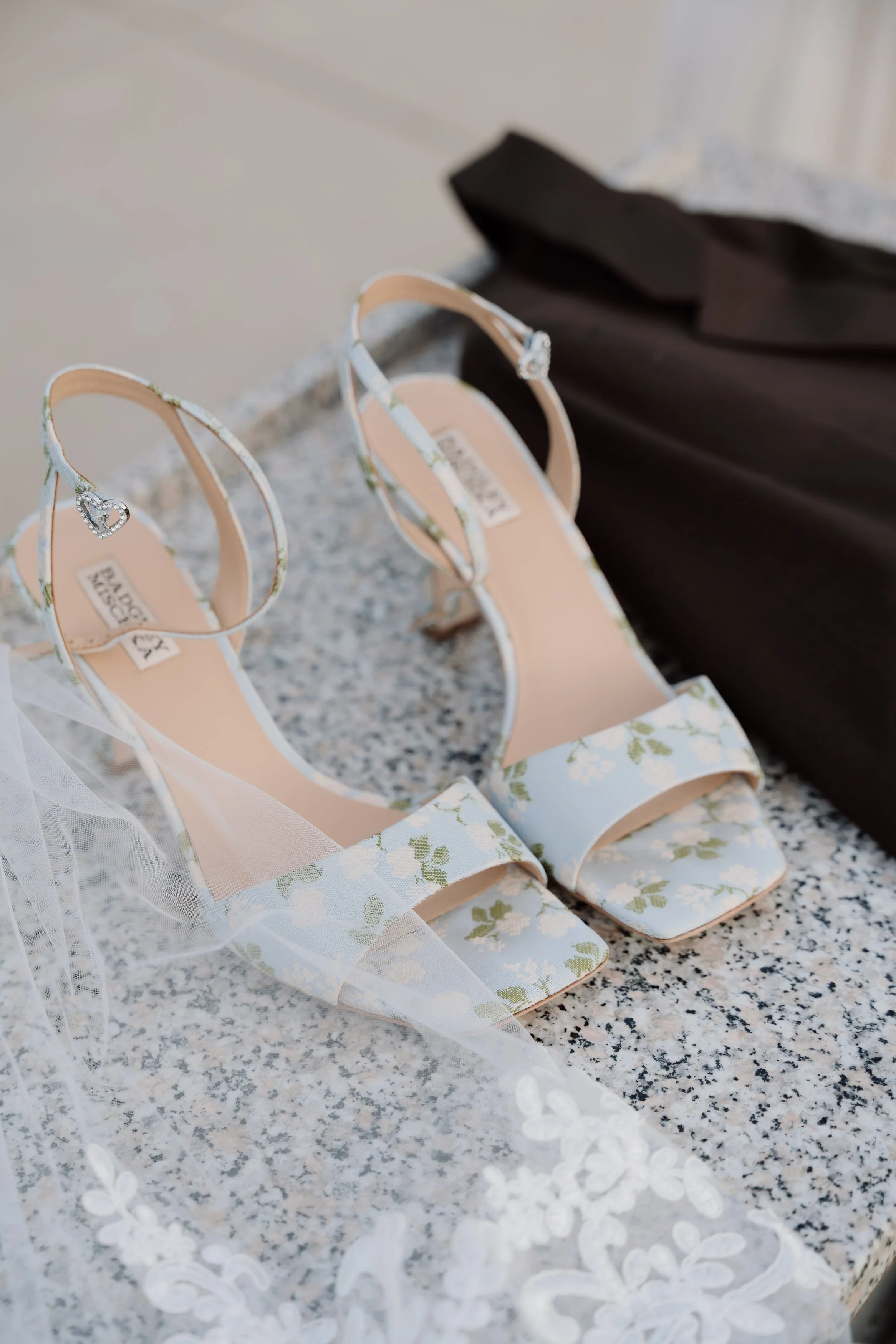 Pair of white floral high-heeled sandals with heart-shaped embellishments and ankle straps, placed on a speckled gray surface. A black garment and white lace fabric are partially visible nearby.