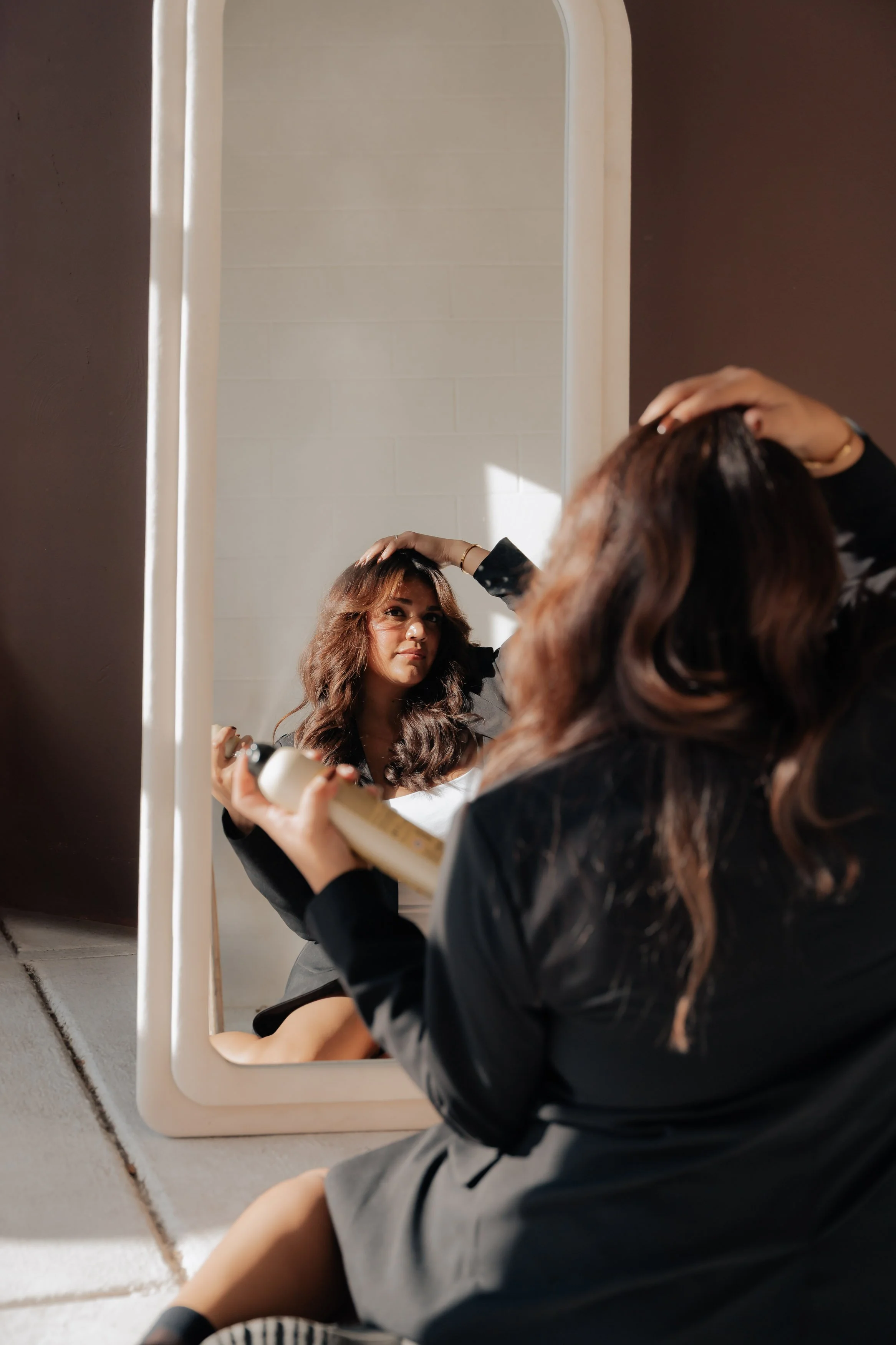 A woman with wavy brown hair sitting on the floor in front of a mirror, using a hair spray, adjusting her hair with one hand, wearing a dark blazer and skirt, and looking at her reflection.