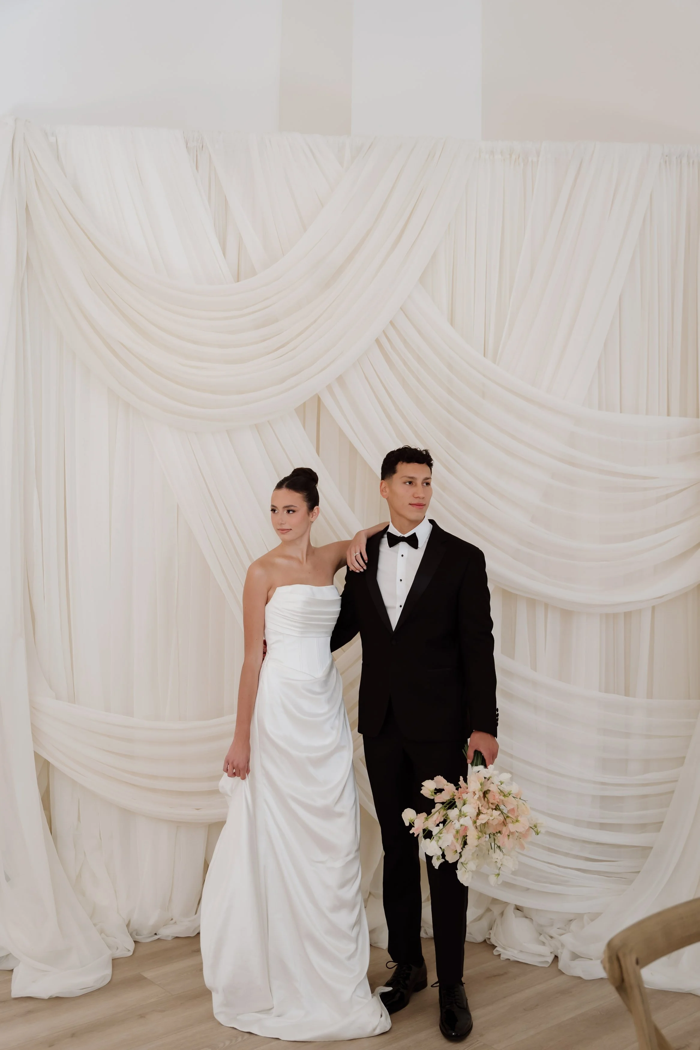 A bride and groom standing in front of a white wedding backdrop with draped fabric. The bride is wearing a strapless white wedding gown, and the groom is dressed in a black tuxedo with a bow tie, holding a bouquet of pink and white flowers.