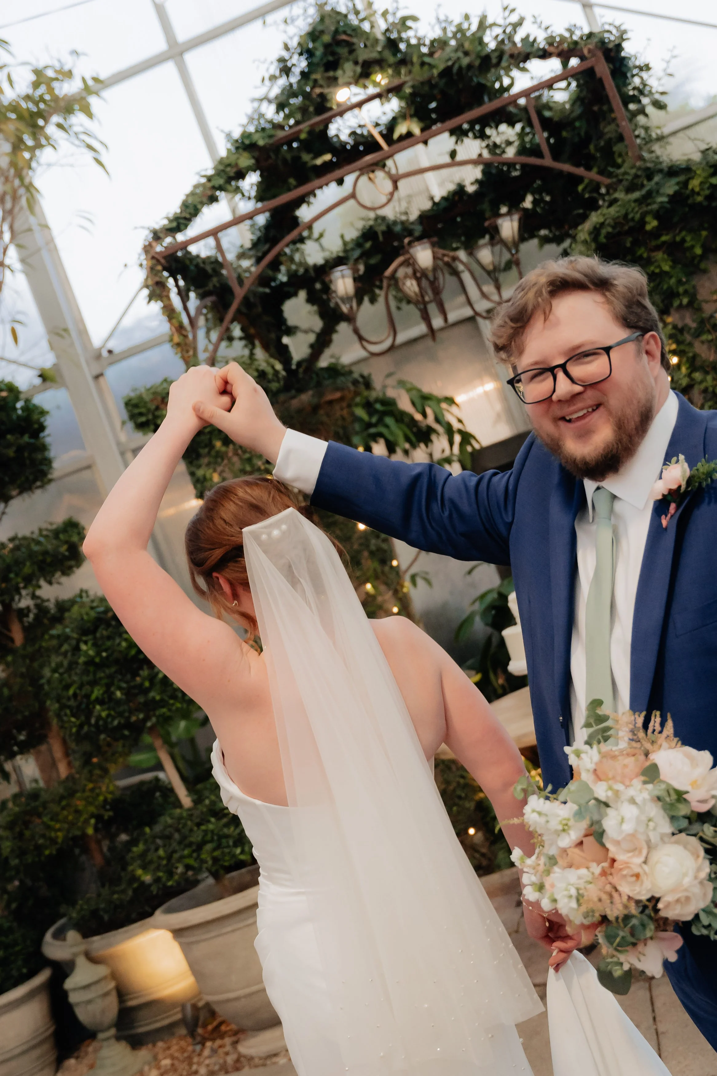 A newlywed couple dancing at their wedding reception, with the groom smiling and wearing glasses and a blue suit, and the bride wearing a white wedding dress and veil, holding a bouquet of light pink and white flowers, inside a greenhouse or garden v