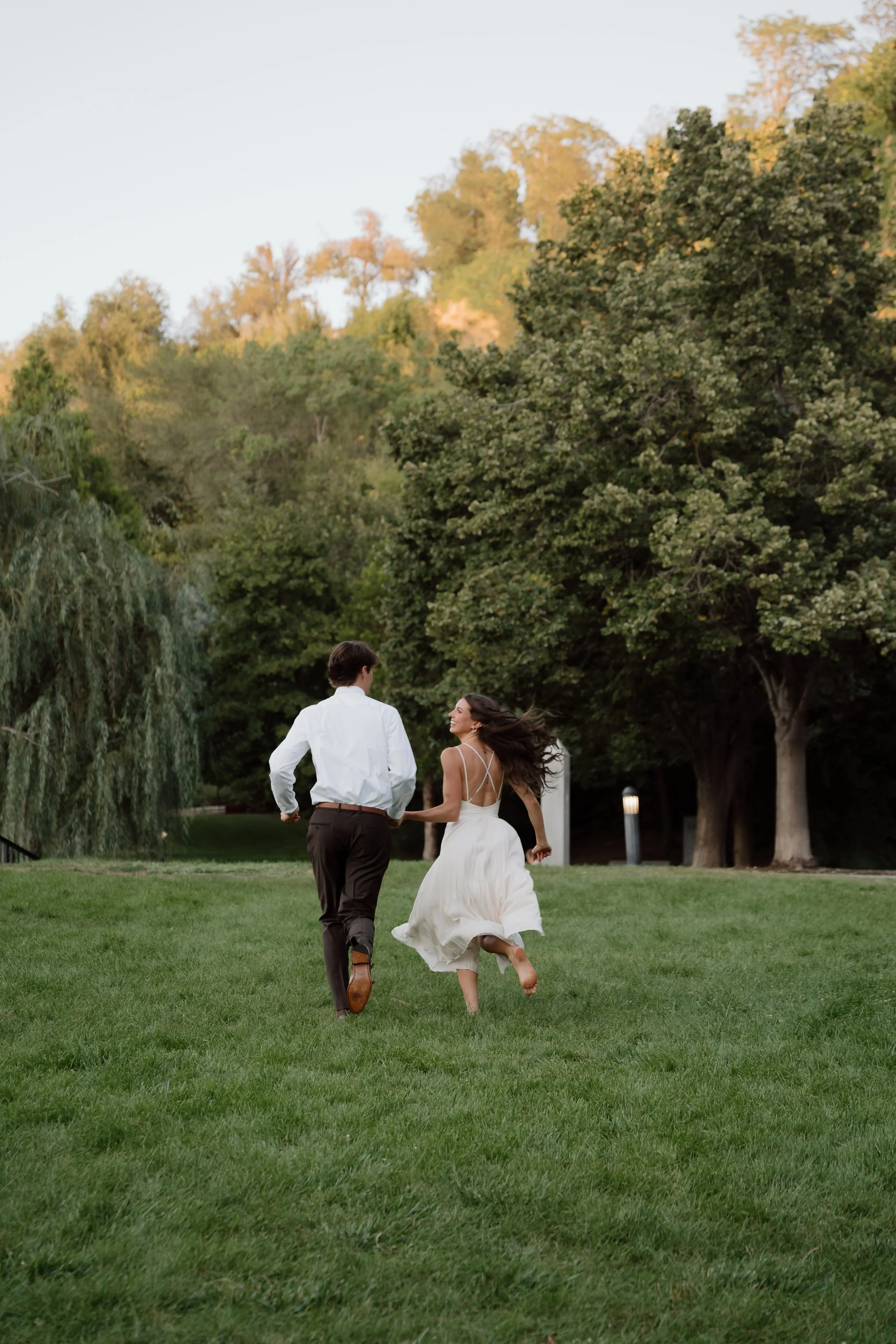 A couple running together in a park, with the woman in a white dress and the man in a white shirt and dark pants, surrounded by green grass and tall trees, during daytime.
