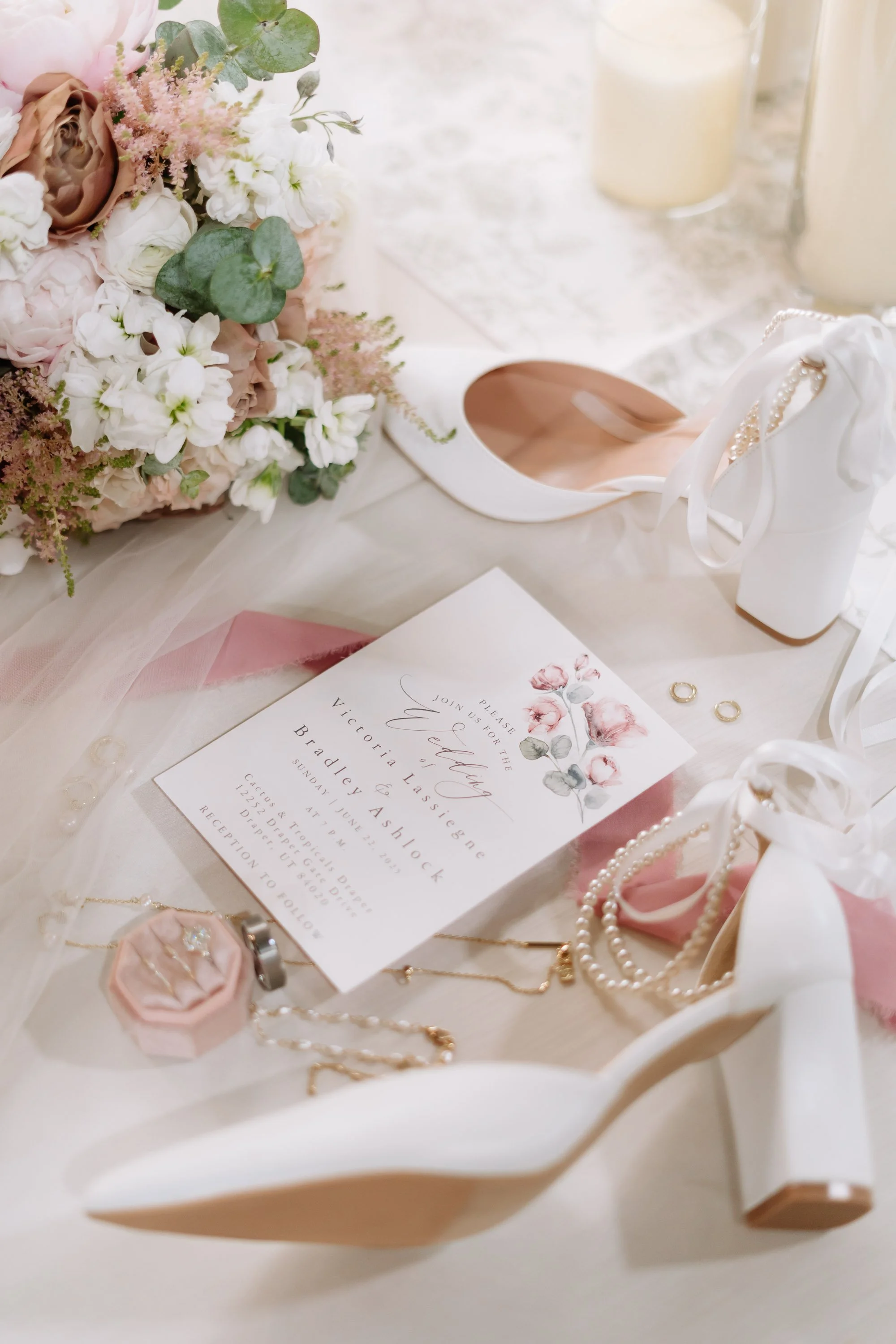 Wedding invitation surrounded by white high heel shoes, pearl necklaces, jewelry, pink velvet ring box, bouquet of pink and white flowers with greenery, and candles.