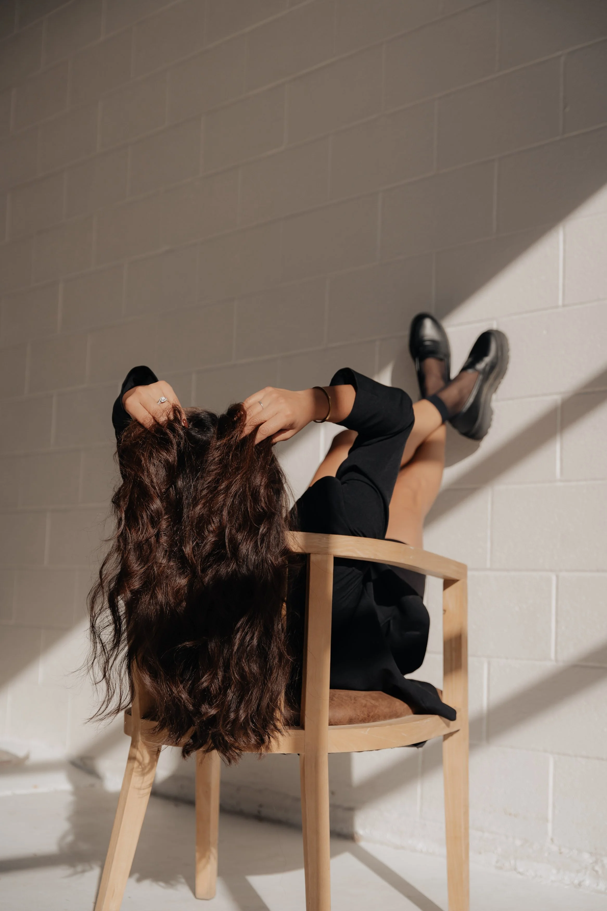 A woman with long, wavy brown hair is sitting on a wooden chair, leaning back with her legs up against a white brick wall, and holding her hair with her hands.