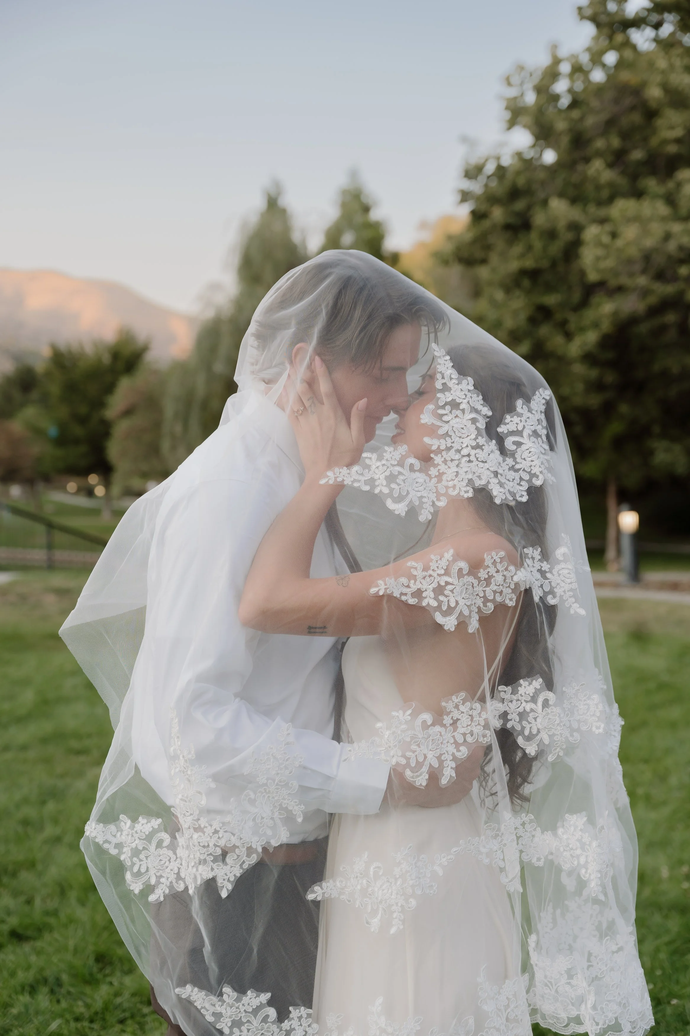 A couple sharing a close, intimate moment outdoors, with the woman wearing a lace veil and dress, and the man holding her face gently.