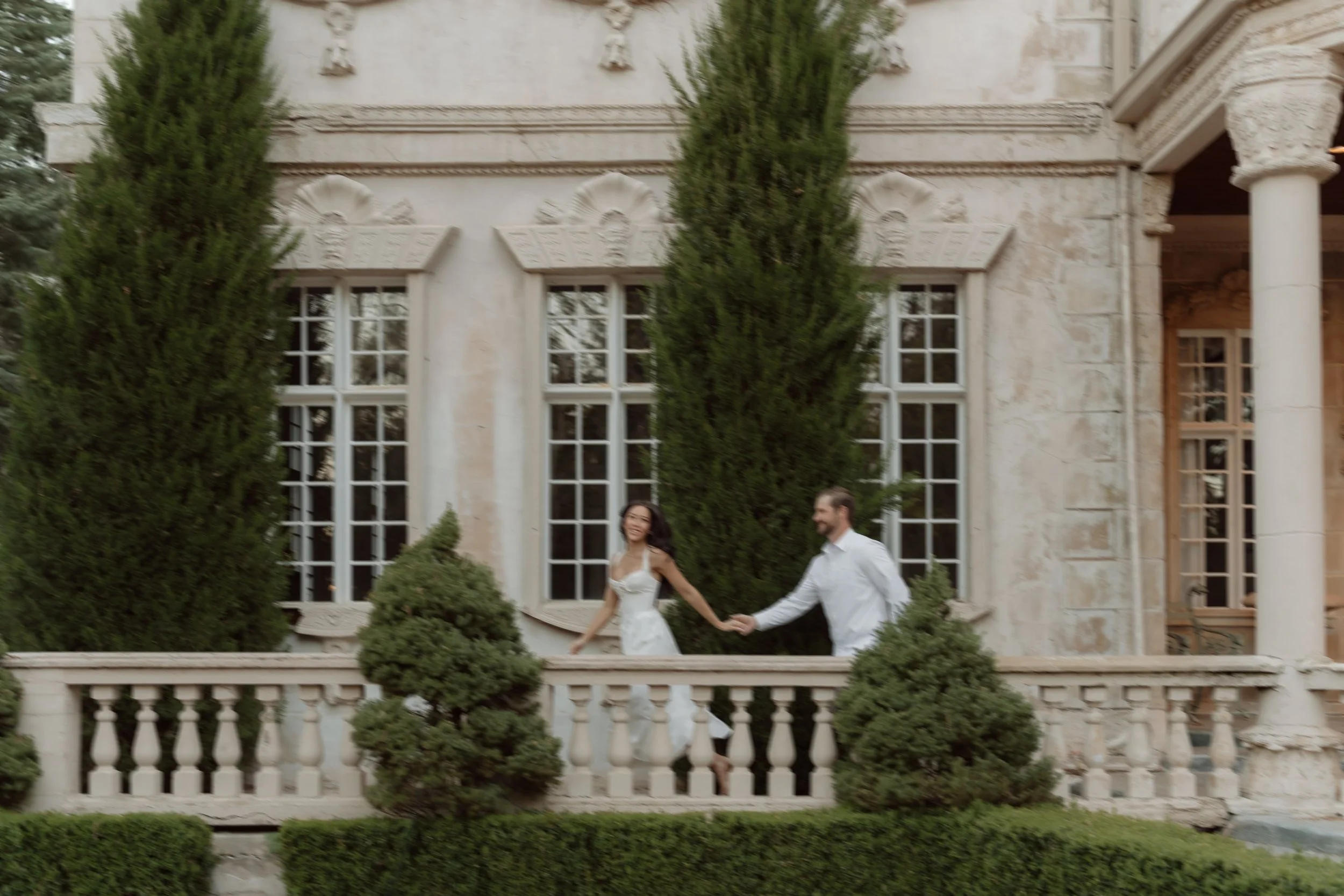 A couple holding hands on a balcony in front of a large ornate building with tall windows and greenery.