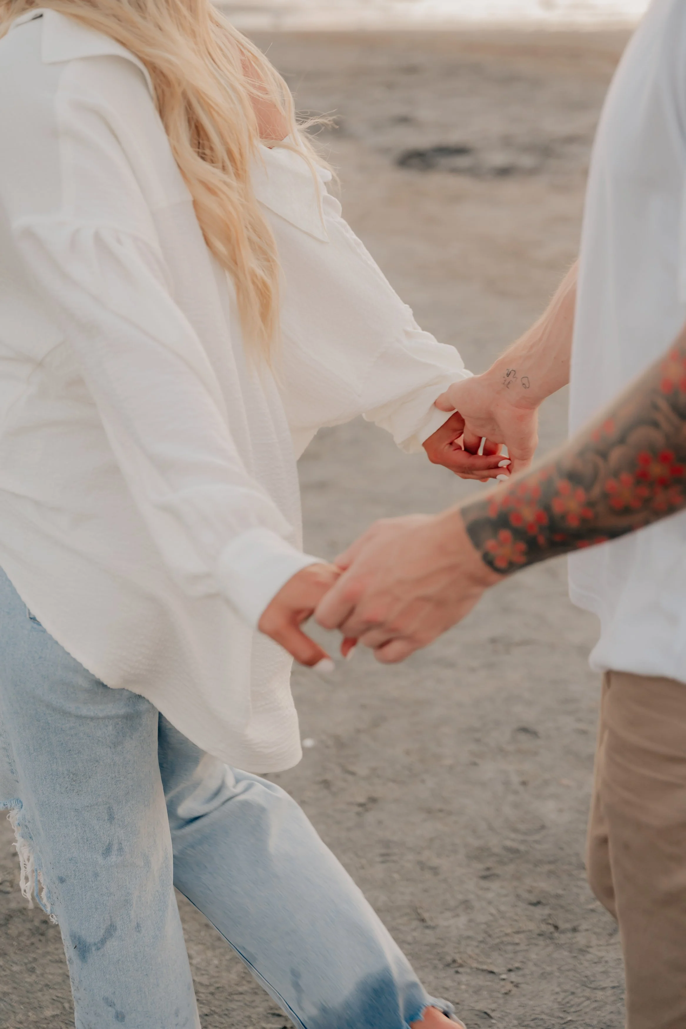 A woman with long blonde hair, wearing a white shirt and light blue ripped jeans, is kneeling on sandy ground holding hands with a man who has a tattooed arm and is wearing beige pants. The background shows a beach or sandy area at sunset.