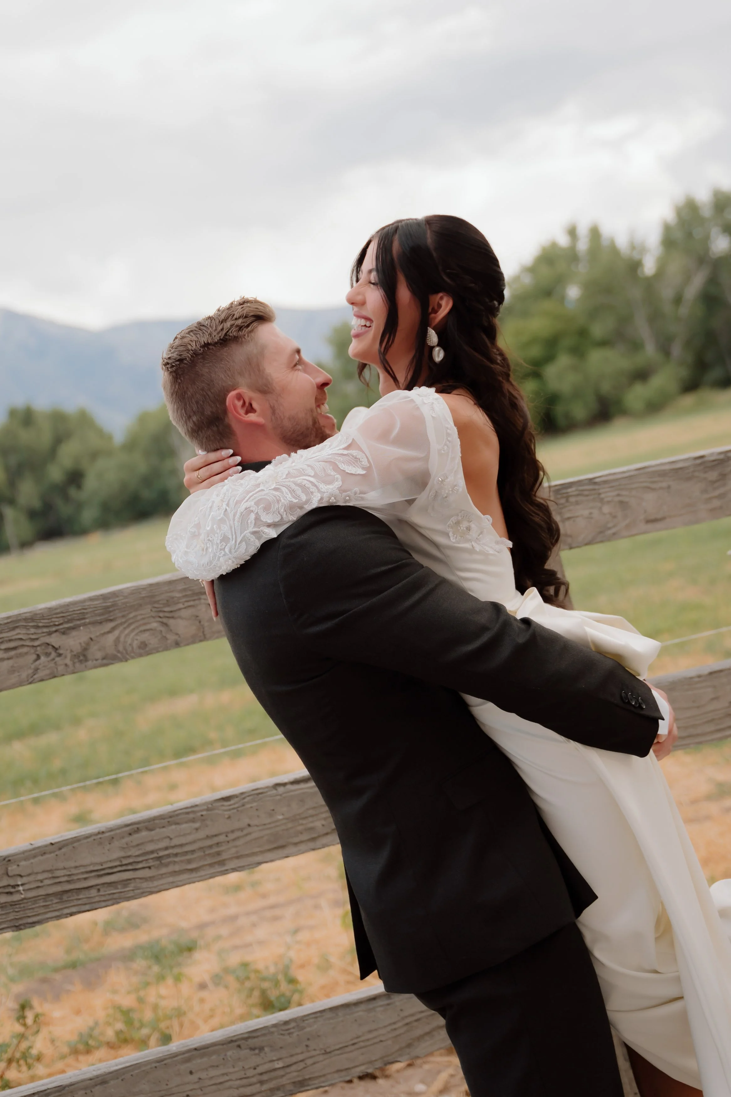 A joyful couple in wedding attire embracing outdoors by a wooden fence, with greenery and mountains in the background.