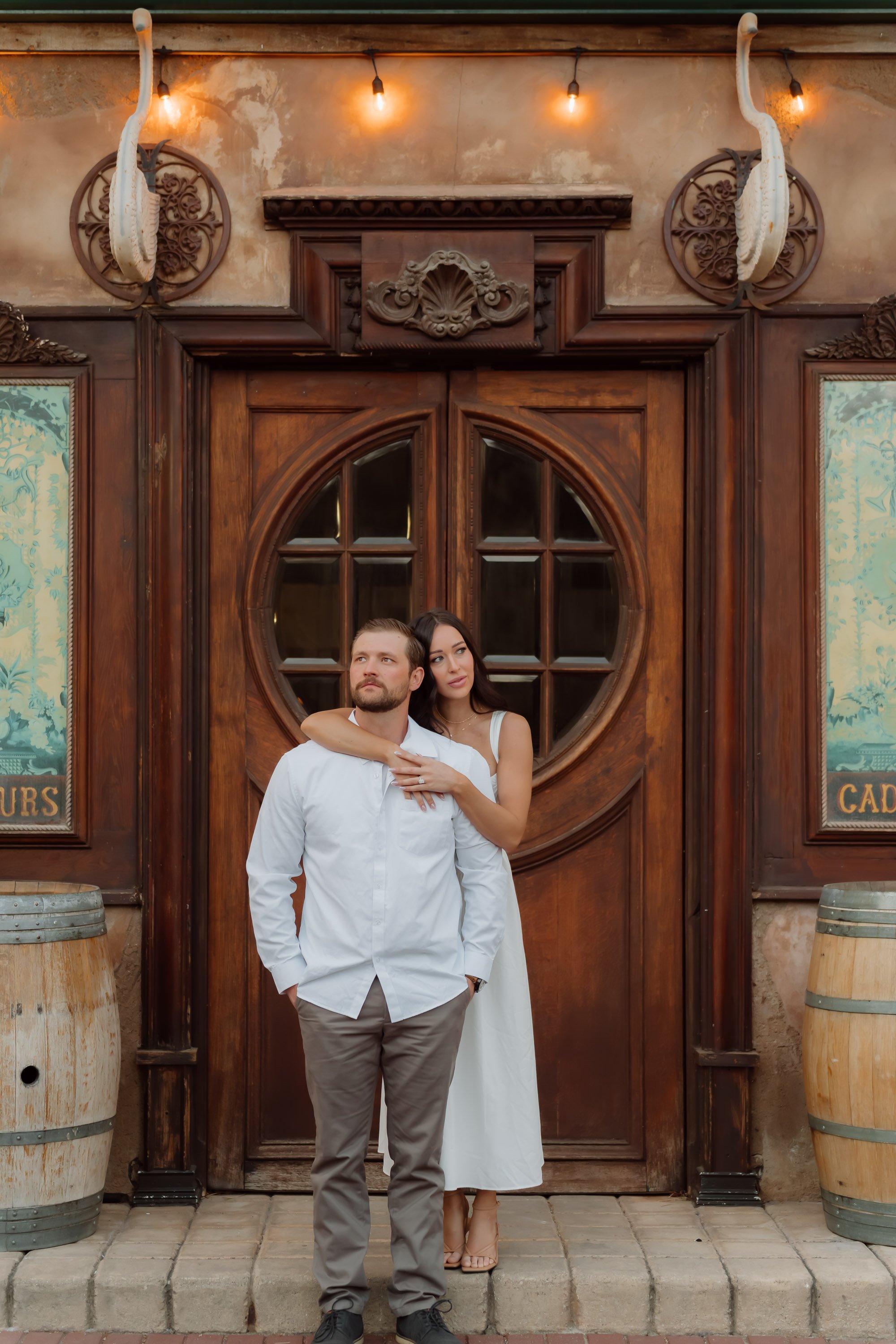 A couple standing in front of a wooden door with decorative carvings. The man is wearing a white shirt and gray pants, with one hand in his pocket. The woman is wearing a white dress, standing behind him with arms around his shoulders. The setting ha