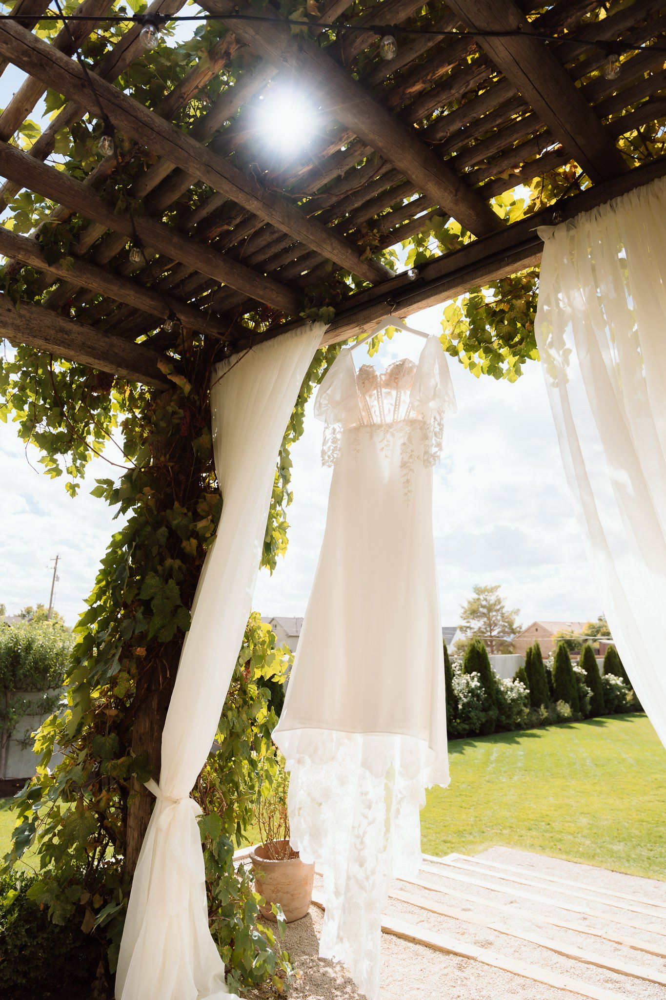 A wedding dress hanging from a wooden pergola adorned with leafy vines and sheer curtains, with bright sunlight and a clear sky in the background.