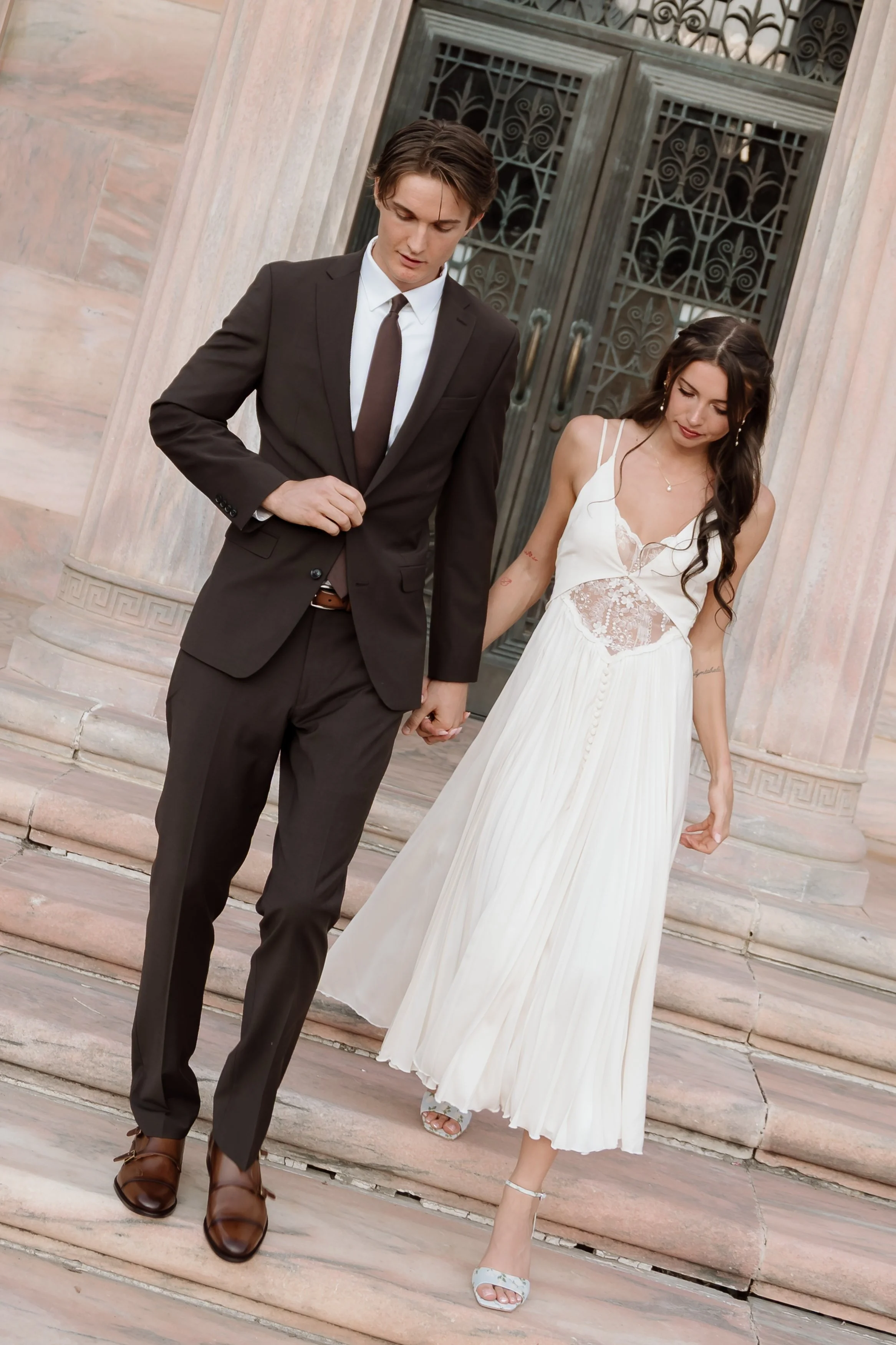 A young couple dressed in formal attire holding hands and descending the steps of a historic building with ornate iron doors.