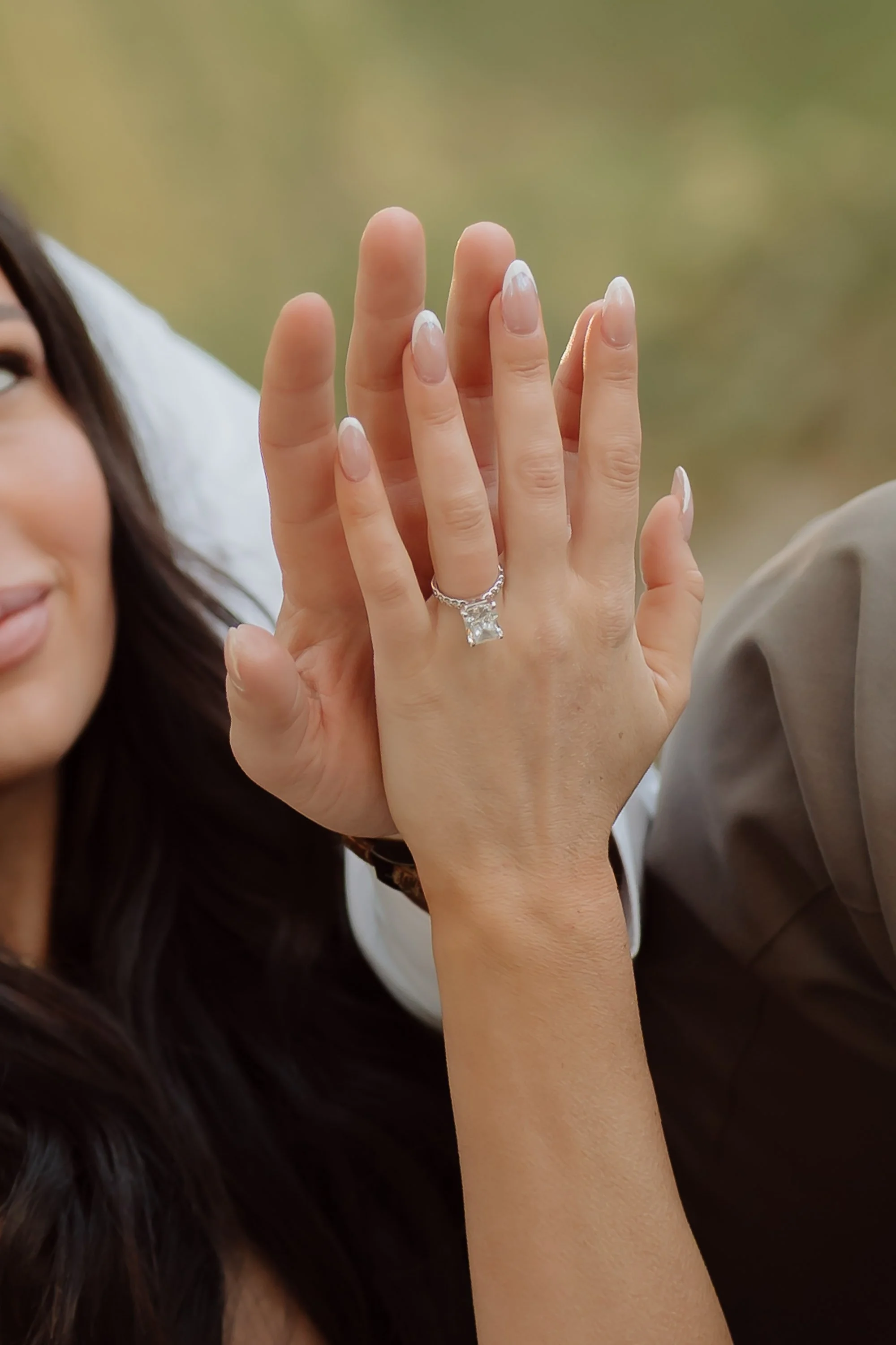 Close-up of a woman's hand with an engagement ring and a man's hand with a wedding band touching together, with part of the woman's face visible on the left side.