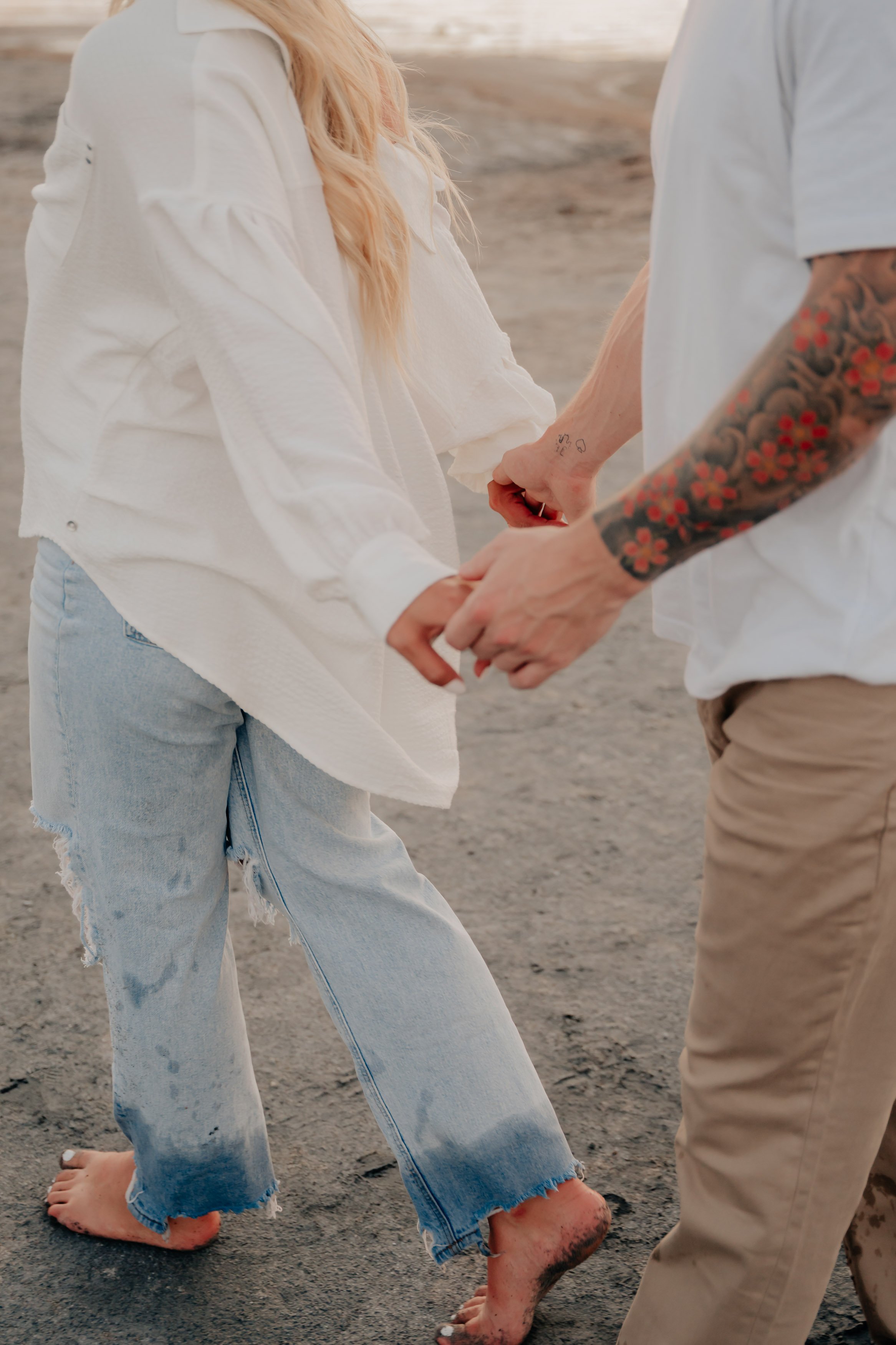 A couple holding hands on a sandy beach with their feet partially covered in wet sand. The woman is barefoot, wearing ripped, light-wash jeans and a white shirt. The man has arm tattoos and is wearing a light gray T-shirt and beige pants.