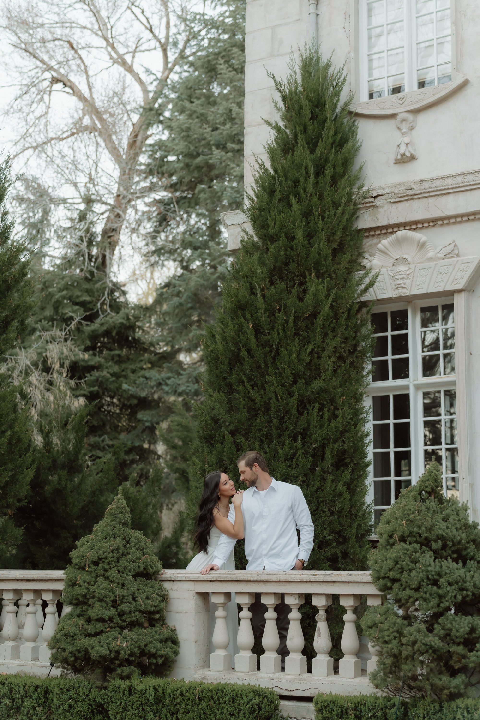 A couple standing on a classical white stone balcony surrounded by lush evergreen trees and foliage, with a historic building featuring ornate decorations and large windows in the background.