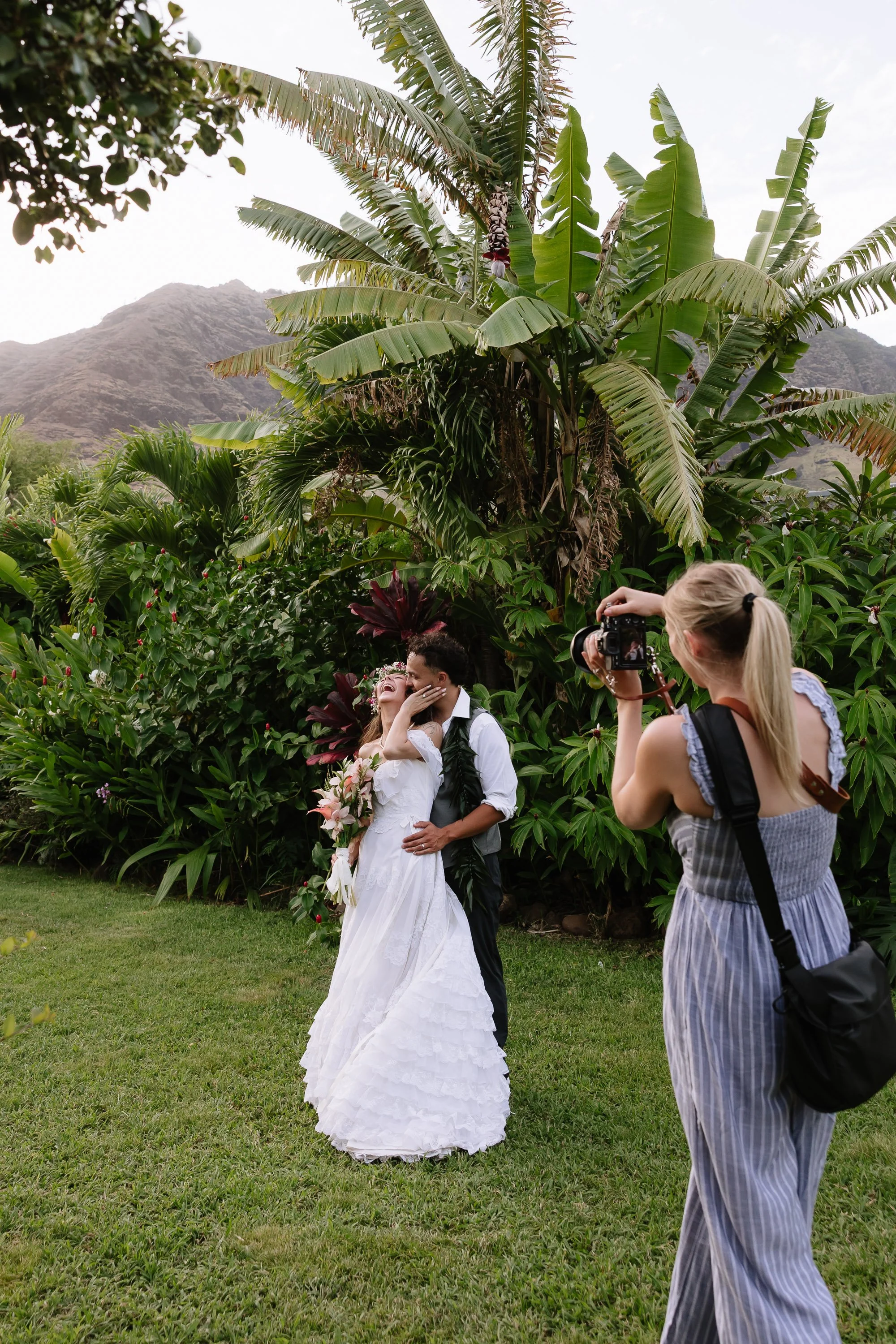 A bride and groom share a romantic moment during their outdoor wedding, with a photographer capturing the scene in a lush, tropical garden.