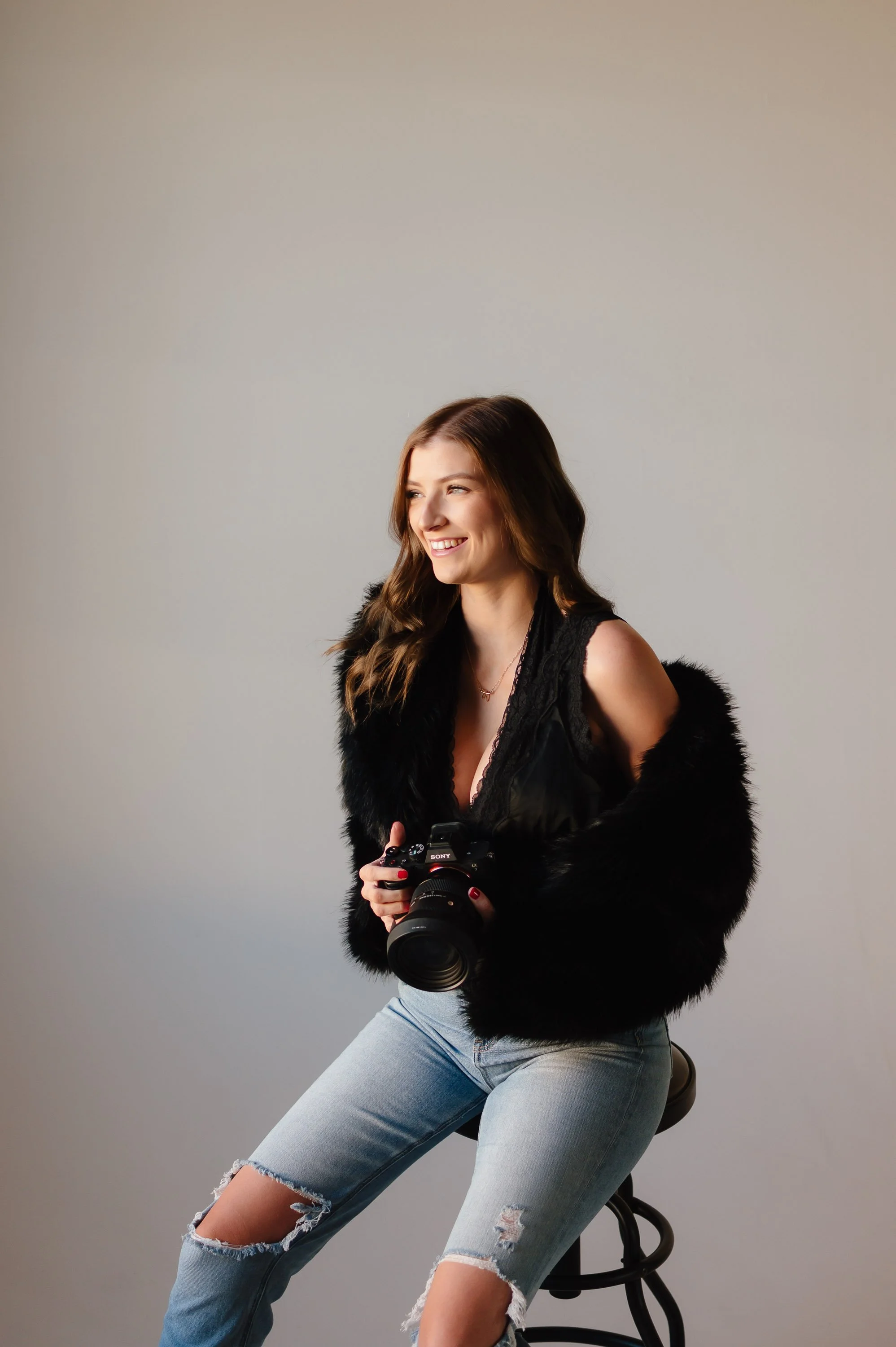 A young woman sitting on a stool, holding a camera, wearing a black sleeveless top with lace details, distressed jeans, and a black fur jacket, smiling against a plain light-colored background.