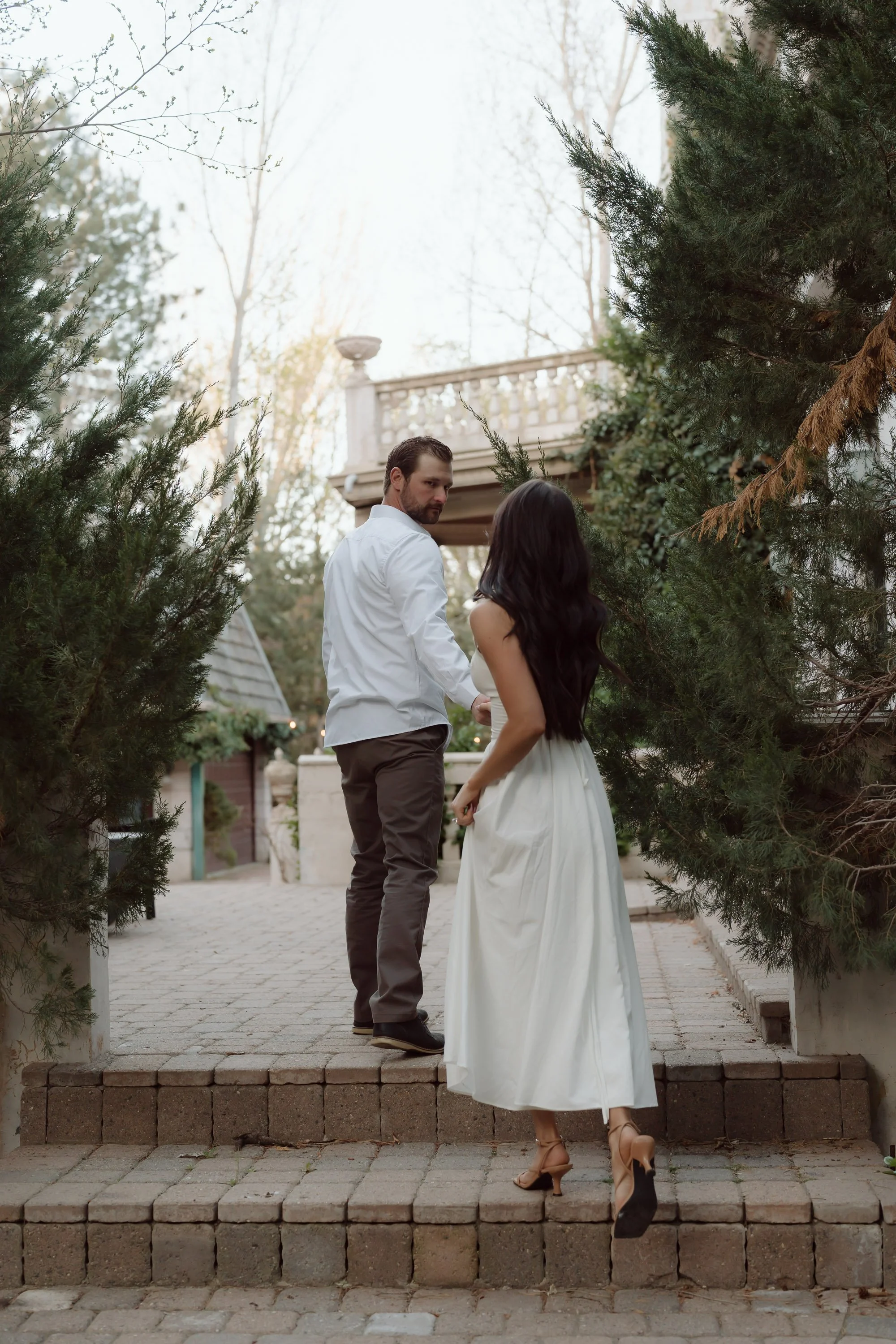 A man and woman holding hands while walking down stone steps outside, surrounded by trees and greenery, with a building structure in the background.