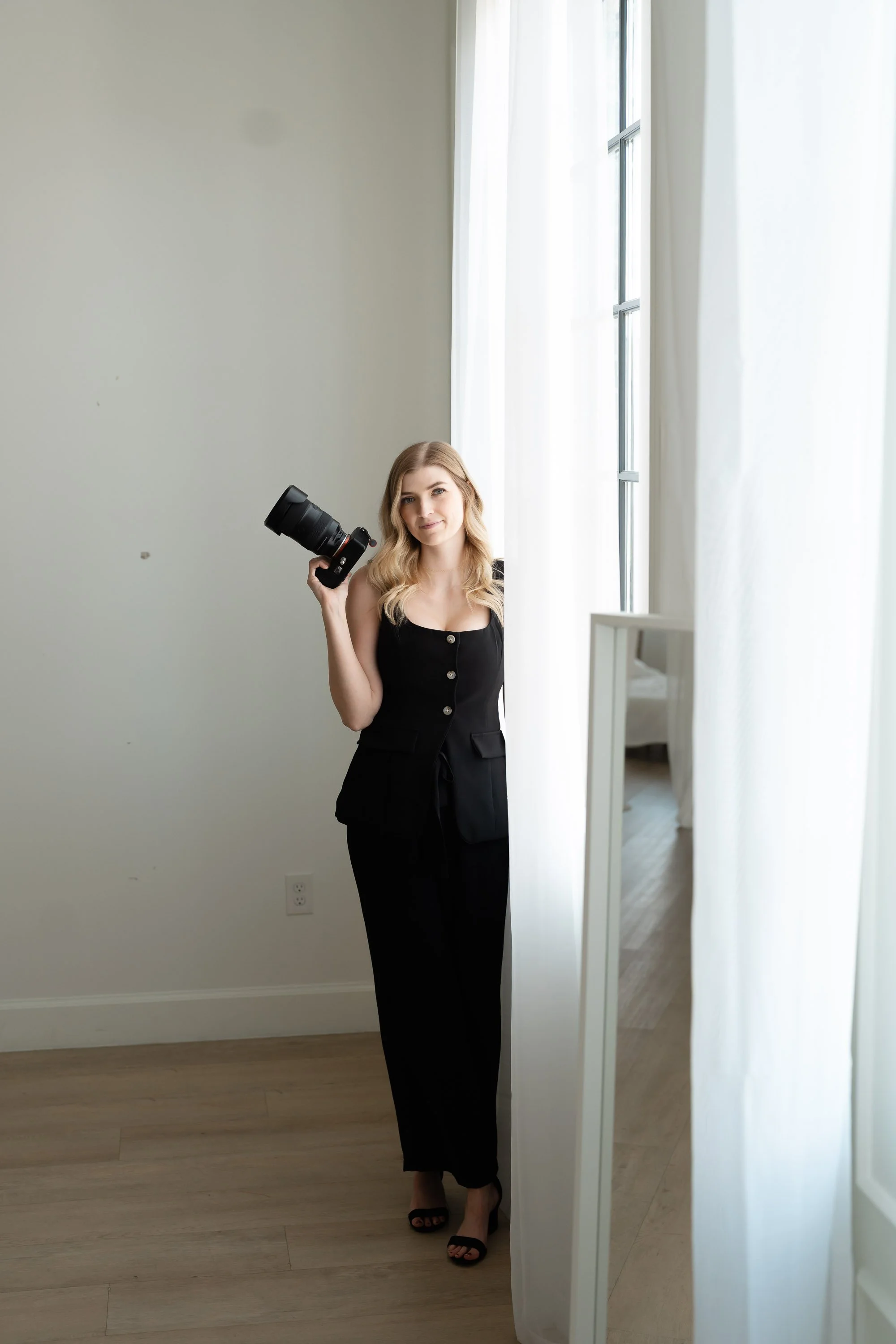 A woman in black pants and sandals standing in front of a large mirror with white curtains, holding a camera, in a room with wooden flooring and plain white walls.