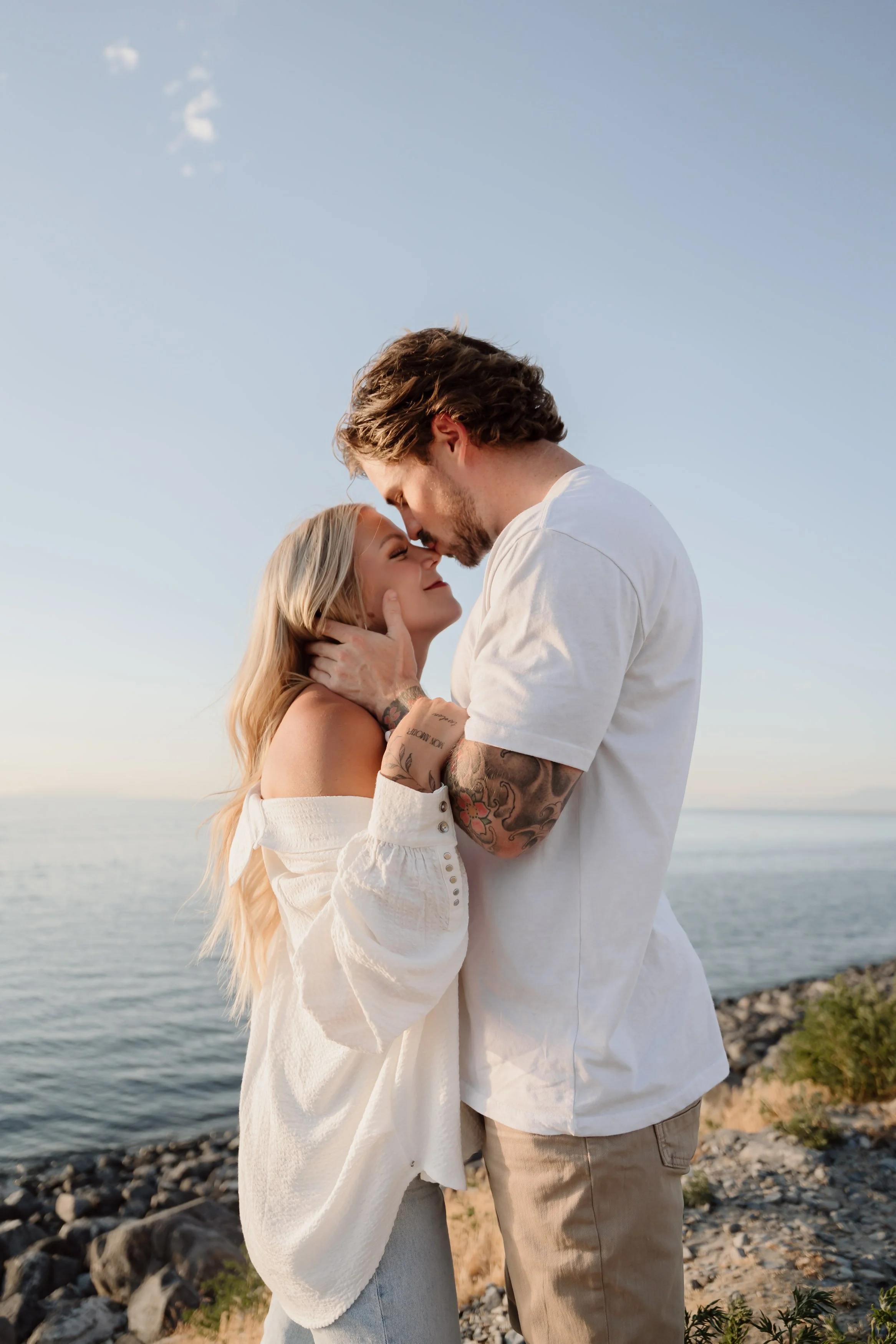A couple embracing on a rocky shoreline, with the ocean and sky in the background, during sunset or early evening.