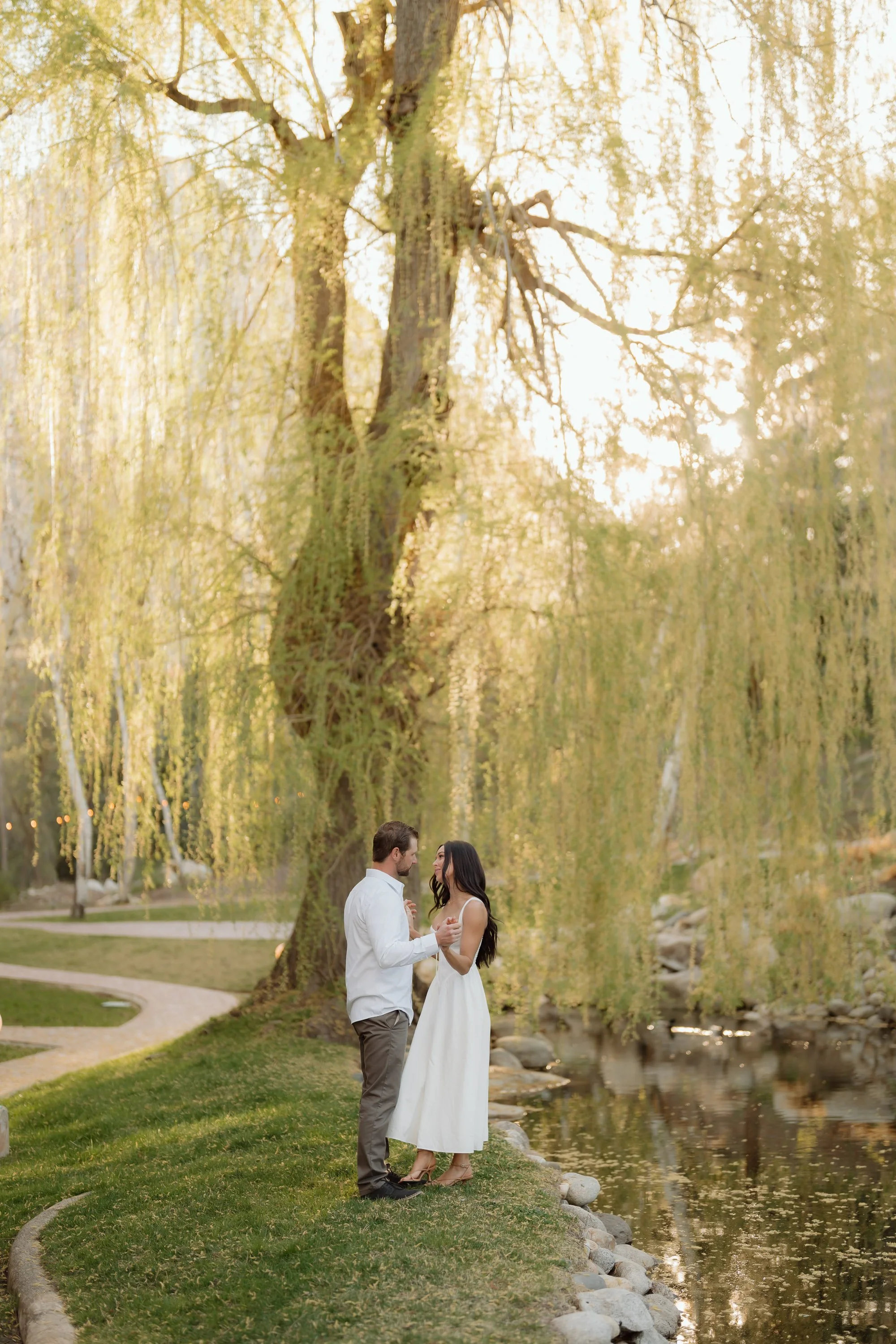 A couple stands close together by a pond in a park during sunset, surrounded by tall trees with green leaves.