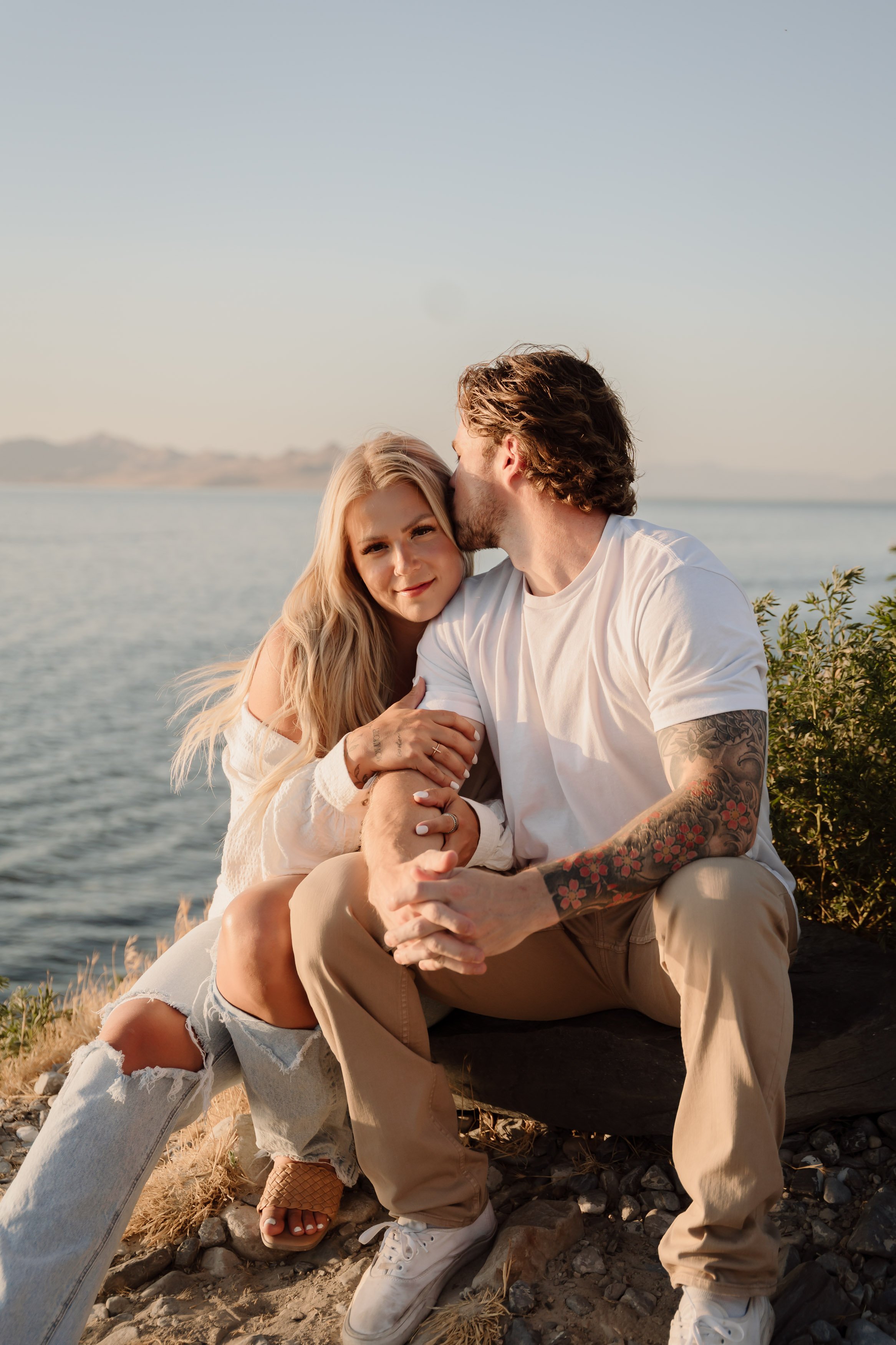 A young couple sitting by the water on a rocky shoreline. The woman has long blonde hair, is smiling softly, and is wearing a white top and ripped light denim jeans. The man has curly brown hair, tattoos on his arms, and is kissing her on the forehea