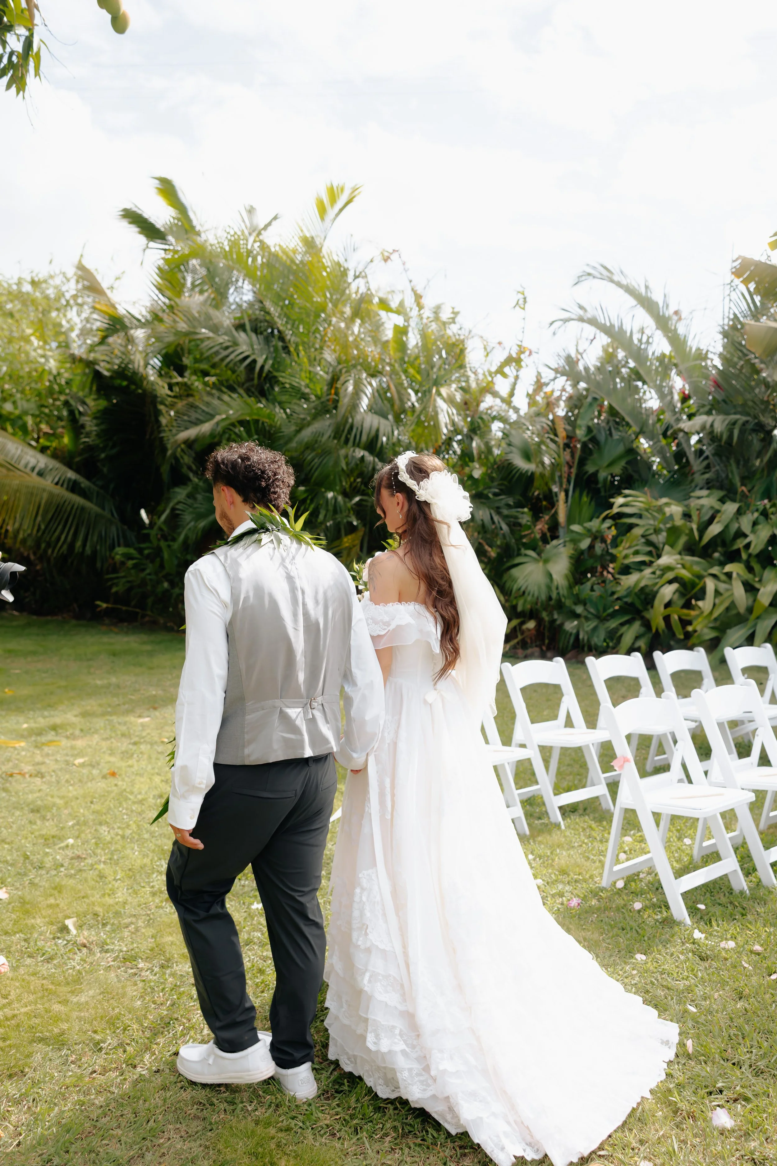 A bride and groom walking hand in hand outdoors at a wedding ceremony, with empty white chairs in the background and lush green tropical foliage beneath a partly cloudy sky.