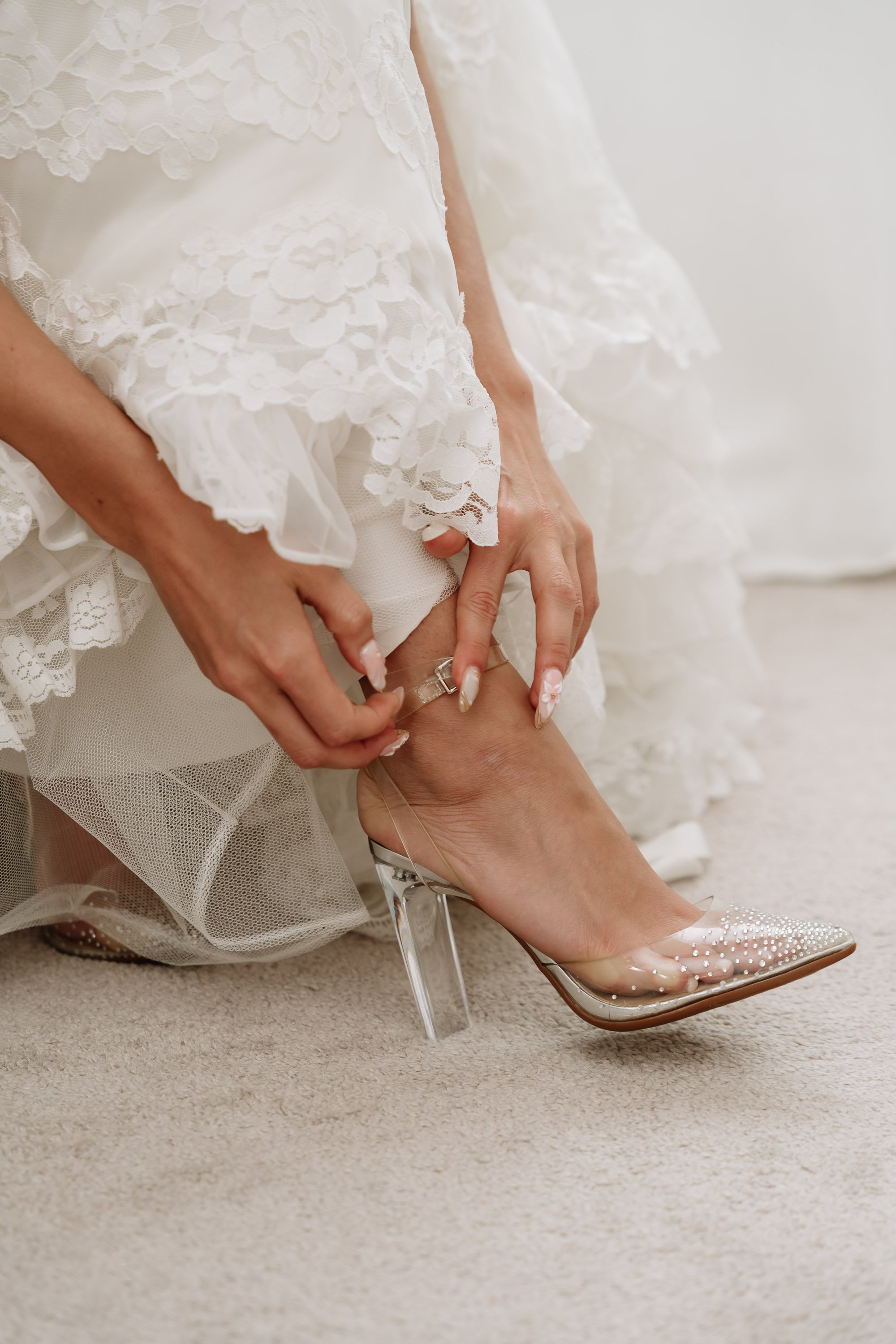 Bridal figure adjusting her elegant high heel with a clear heel and decorated with pearls, while wearing a lace wedding dress.