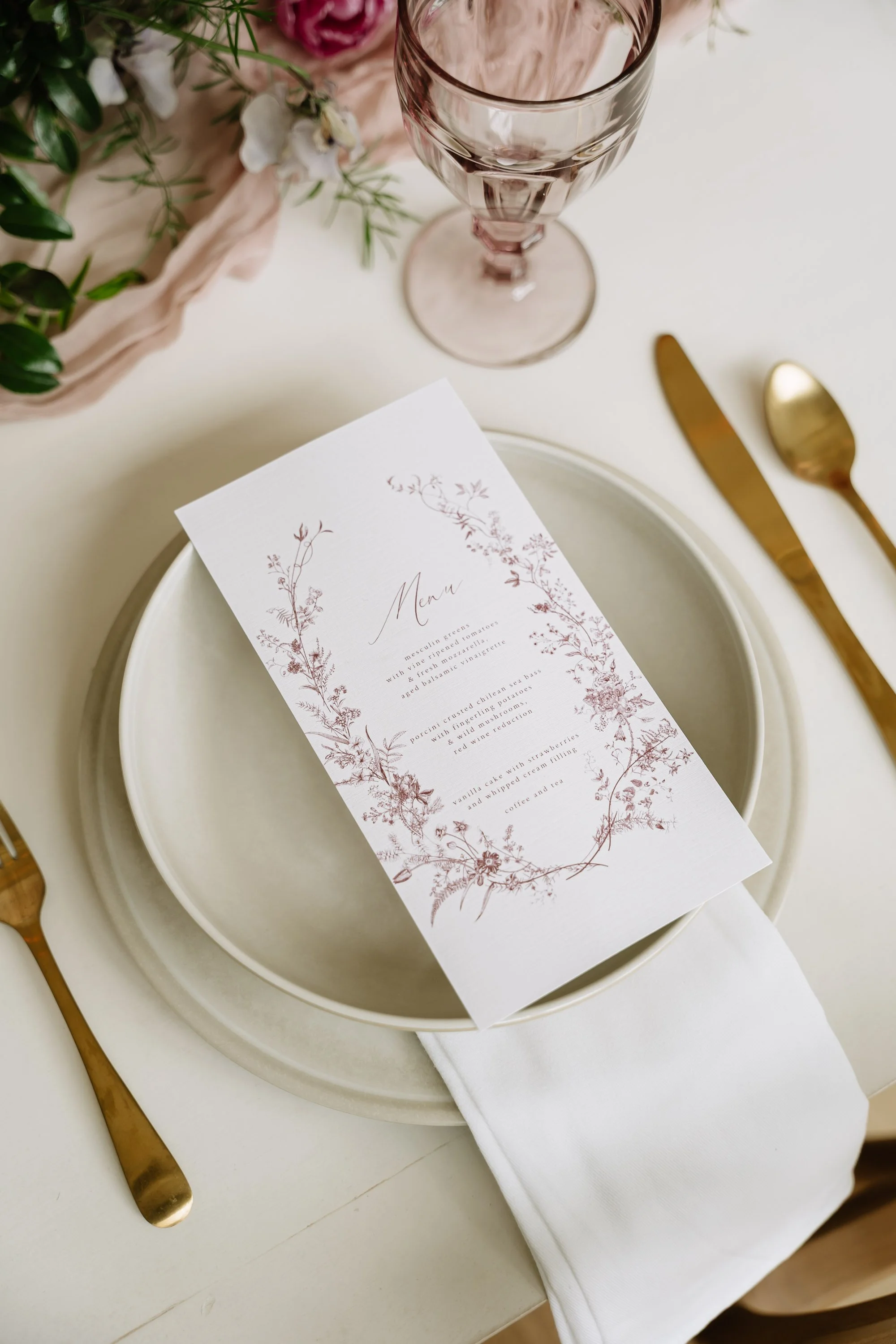 Elegant table setting with a menu card placed on a white plate, gold cutlery, a pink wine glass, and floral decorations in the background.