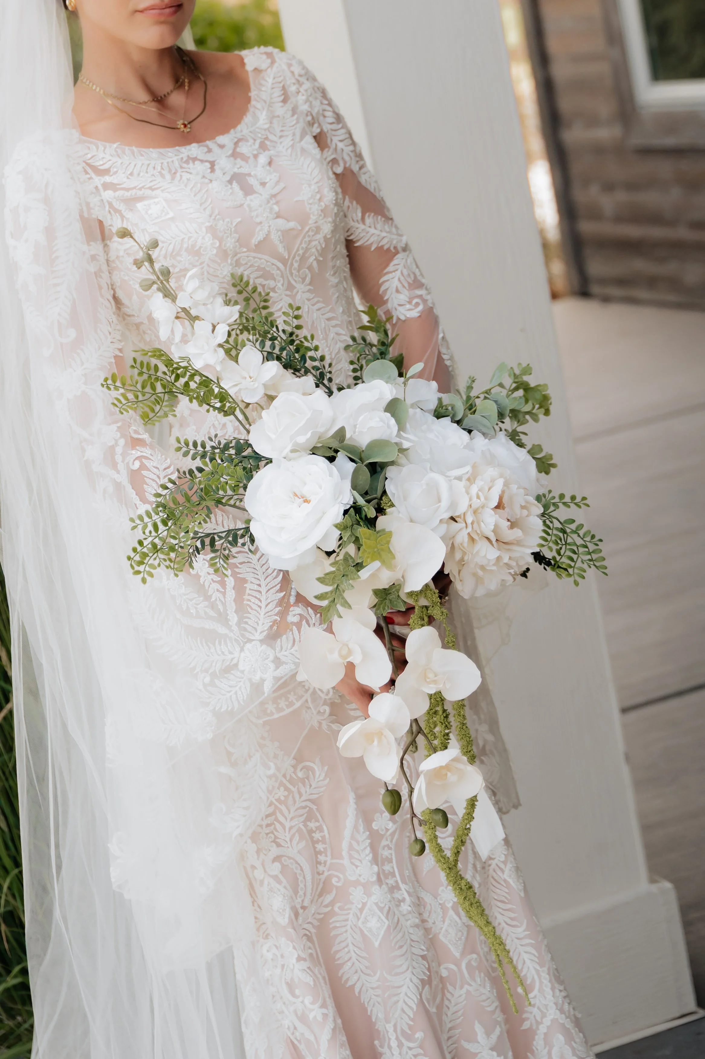 Bride holding a cascading bouquet of white flowers, including roses and orchids, in an off-white wedding gown with lace embroidery.