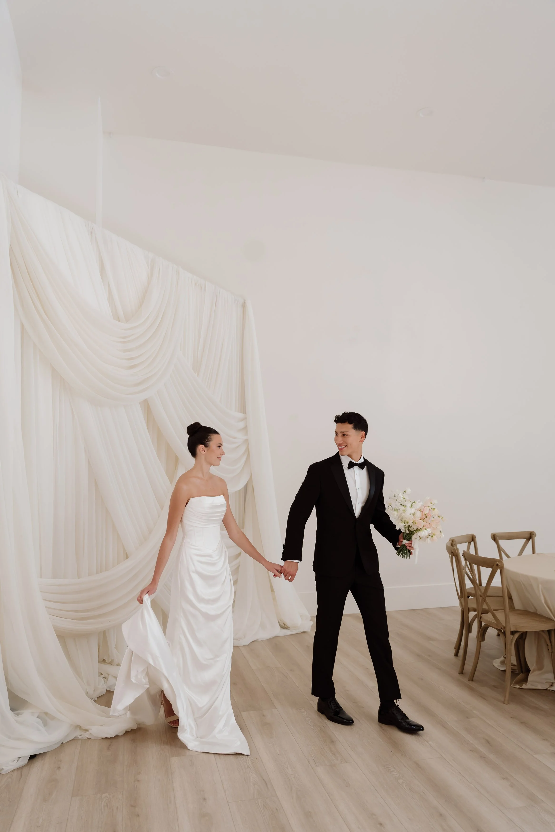 Bride and groom holding hands inside a minimalist wedding venue, with white draped fabric as a backdrop and wooden chairs and table to the side.