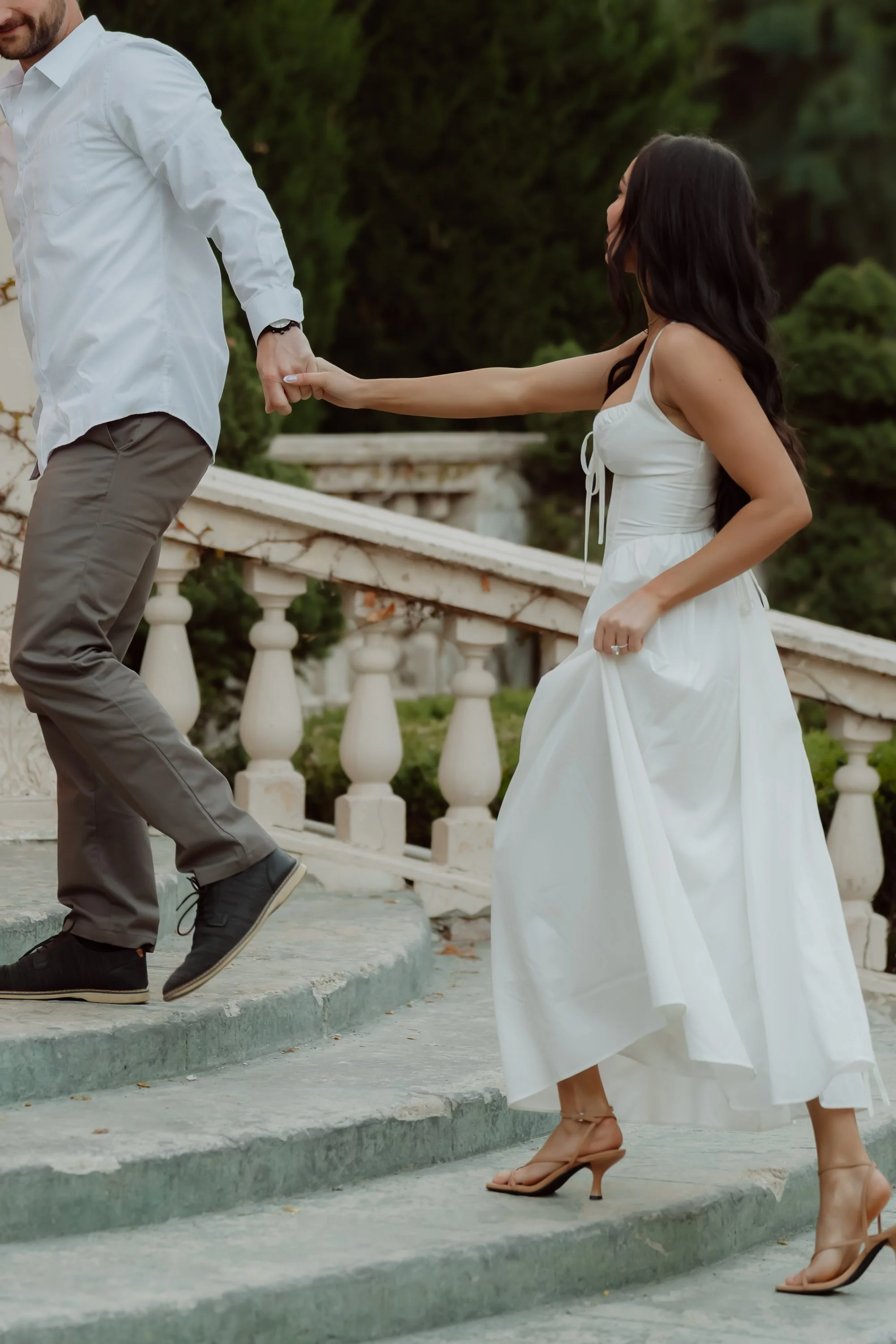 A woman in a white dress helping a man in a white shirt and brown pants ascend stairs outdoors. The woman is holding the man's hand and is mid-step on the stairs, wearing beige high-heeled sandals with ankle straps. The background features greenery a