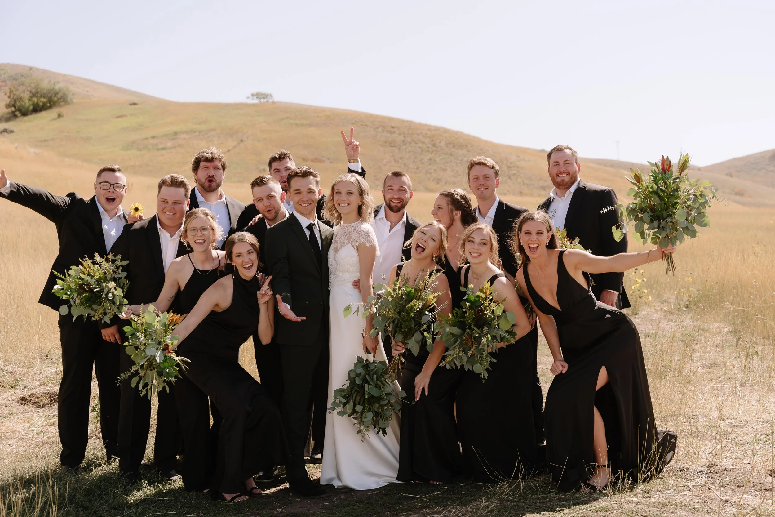 A group of people, including a bride and groom, is celebrating outdoors in a grassy field with hills in the background. They are dressed in formal attire, with the women in black dresses and the men in suits. Some are holding bouquets and all appear happy.