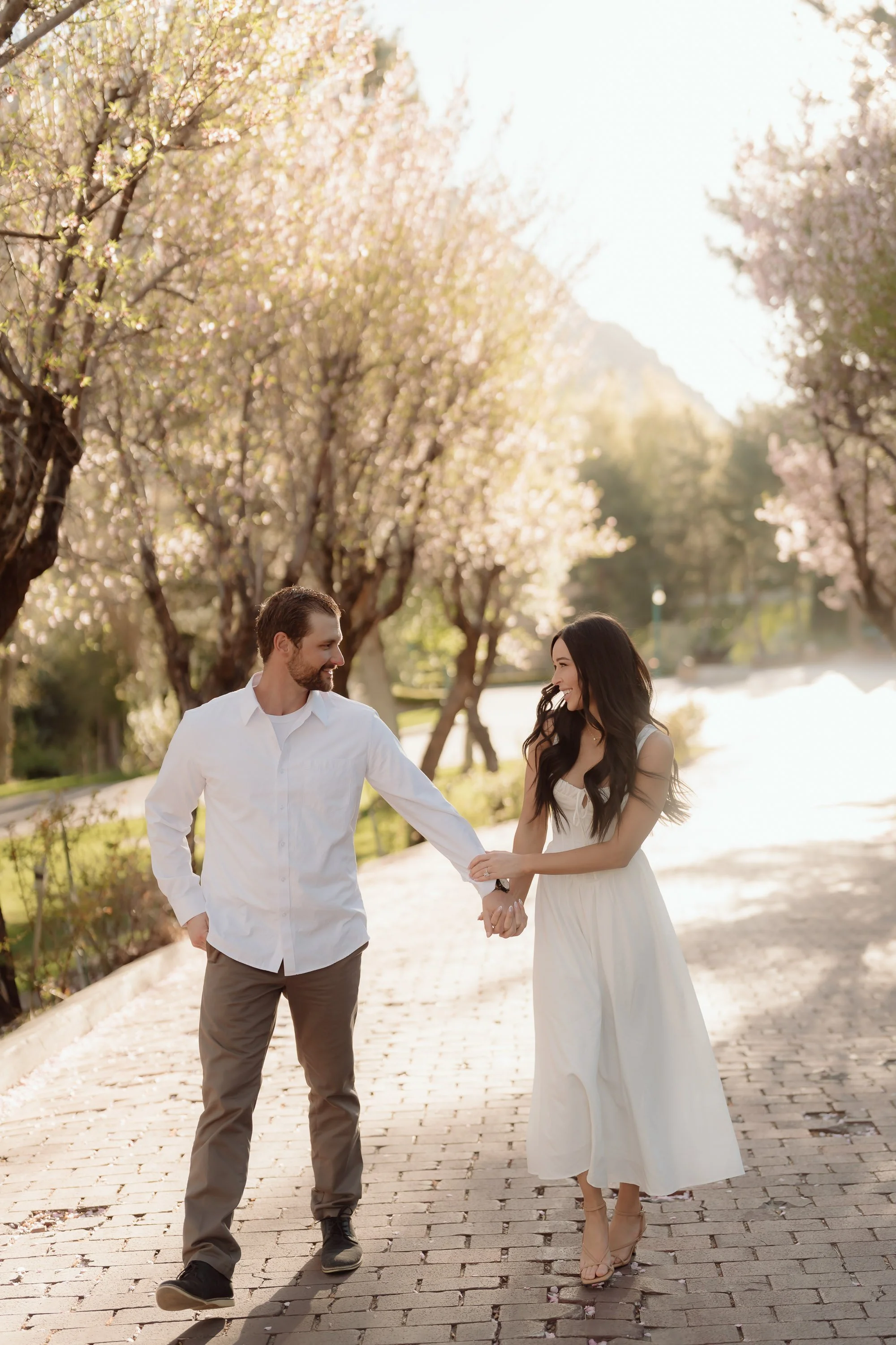 A happy couple standing on a cobblestone path surrounded by pink cherry blossom trees during daytime, holding hands and smiling at each other.