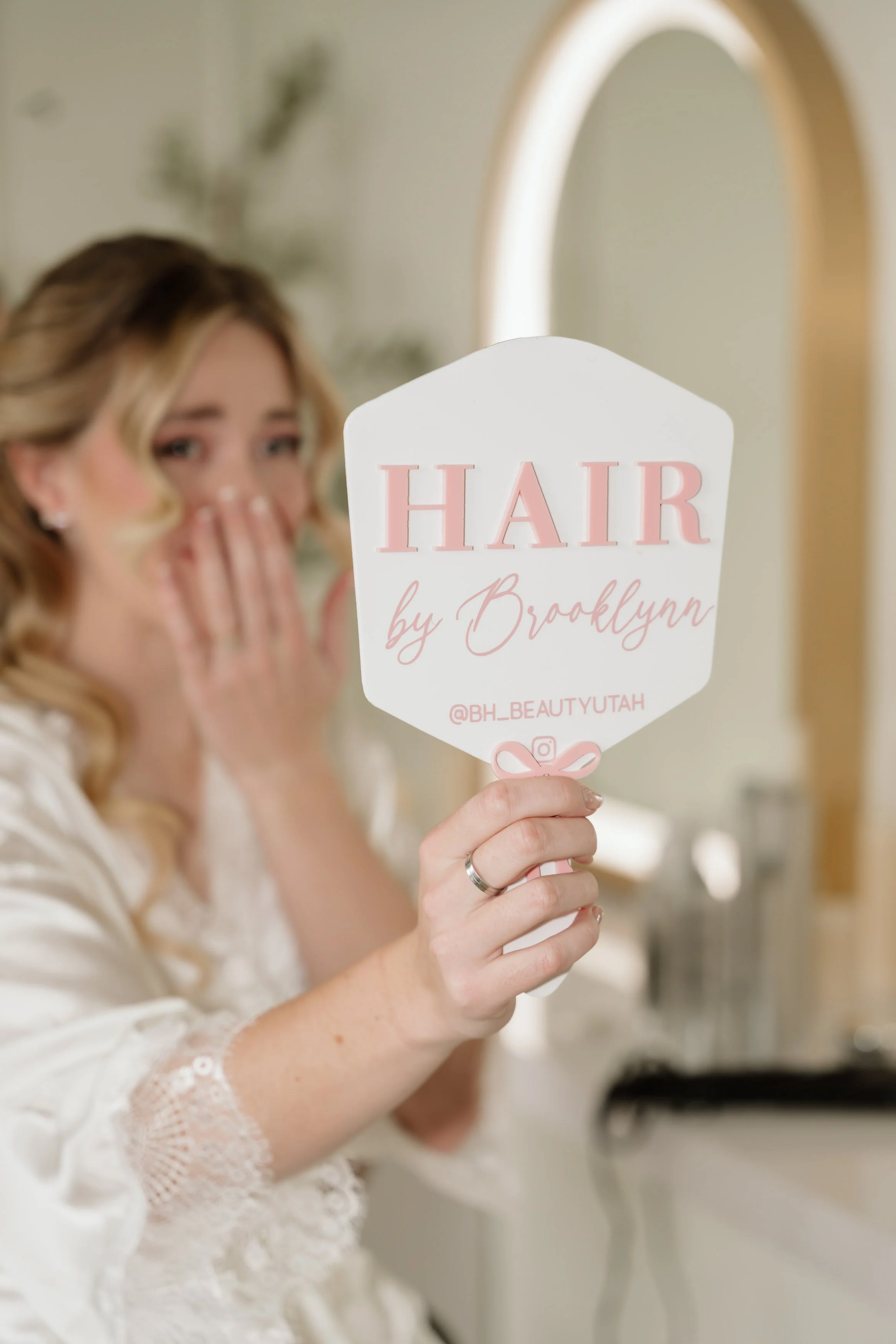 A woman holding a pink and white sign that reads 'HAIR by Brooklyn' with a bow and Instagram handle '@BH_BEAUTYUTAH', while looking in a mirror.