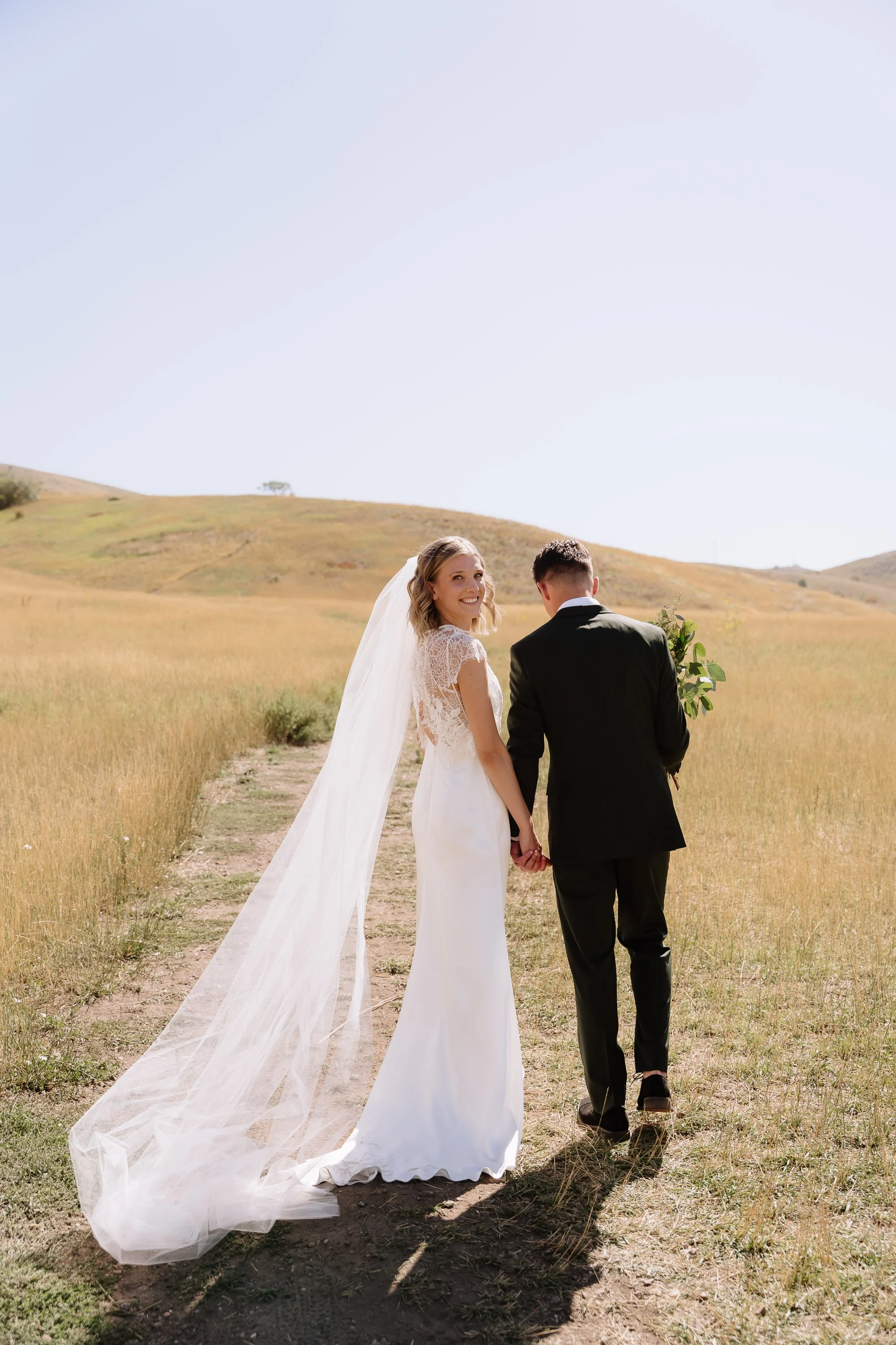 A bride and groom walk hand-in-hand through a grassy field, holding a bouquet, with rolling hills in the background.