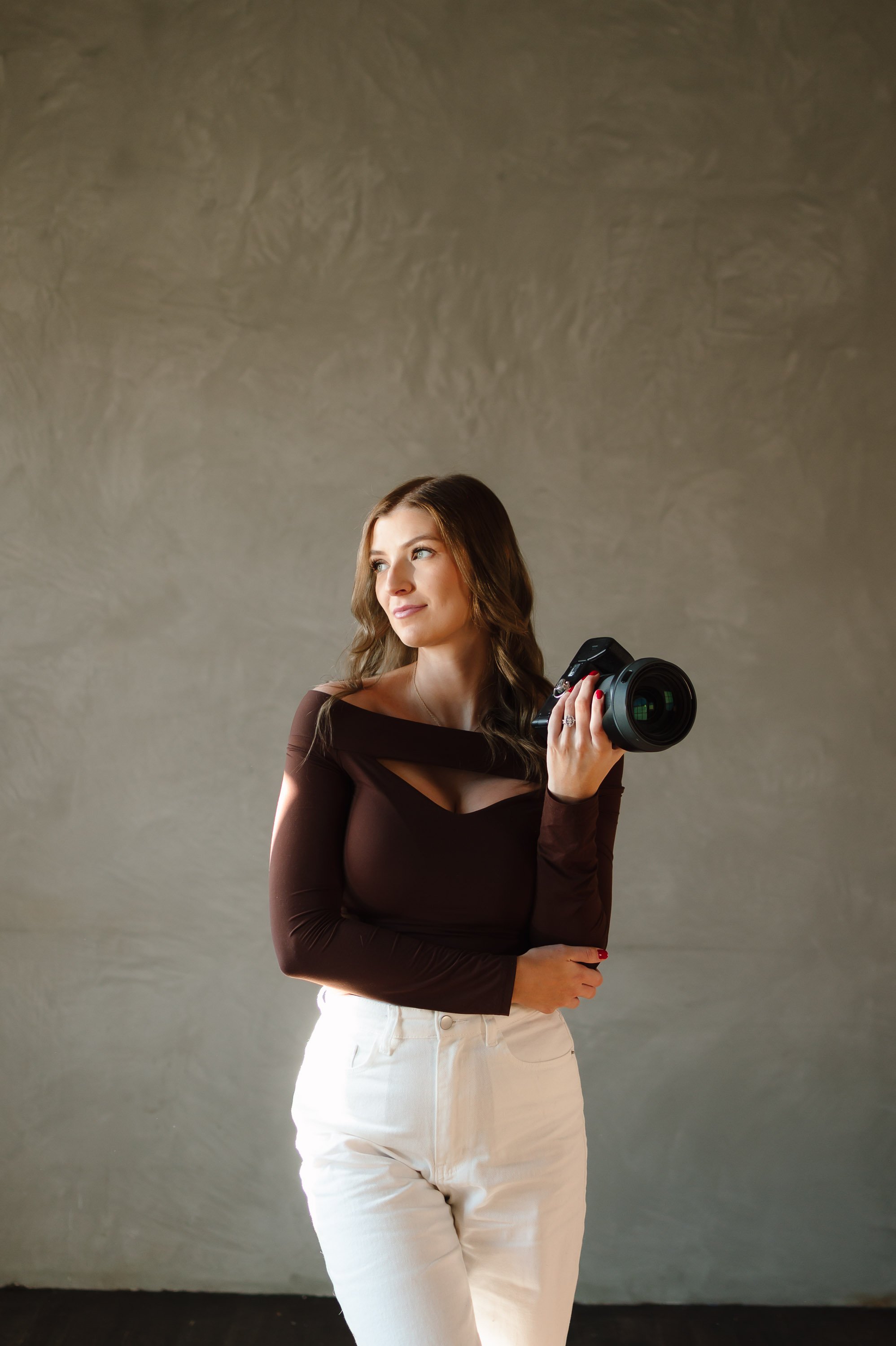 A woman with long brown hair wearing a black top and white pants holding a camera, standing in front of a plain gray background.