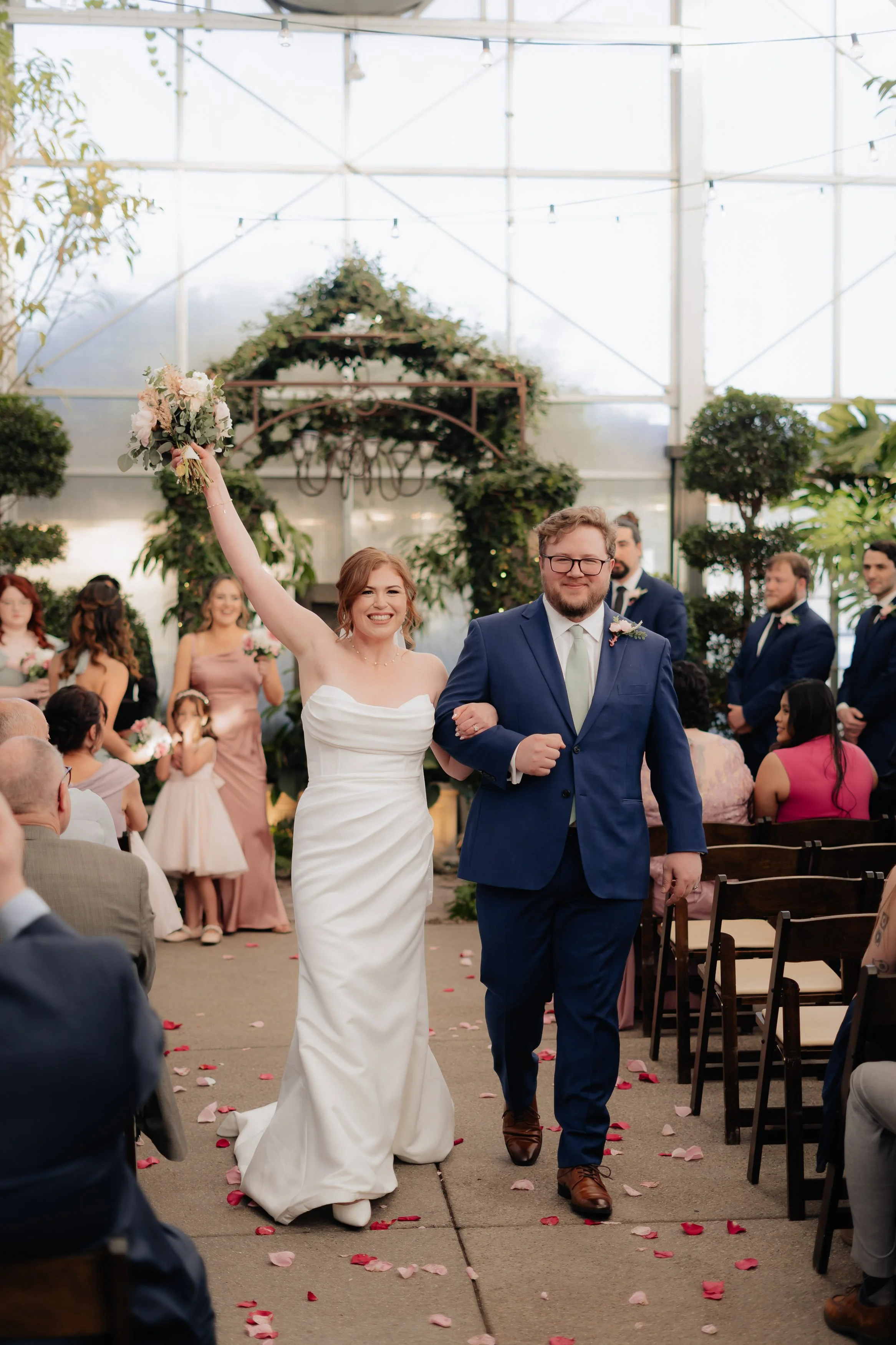 A newlywed couple walking down the aisle after wedding ceremony in a glass-ceiling venue, surrounded by friends and family, with guests seated on either side, flower petals scattered on the ground, and wedding party members standing in the background.