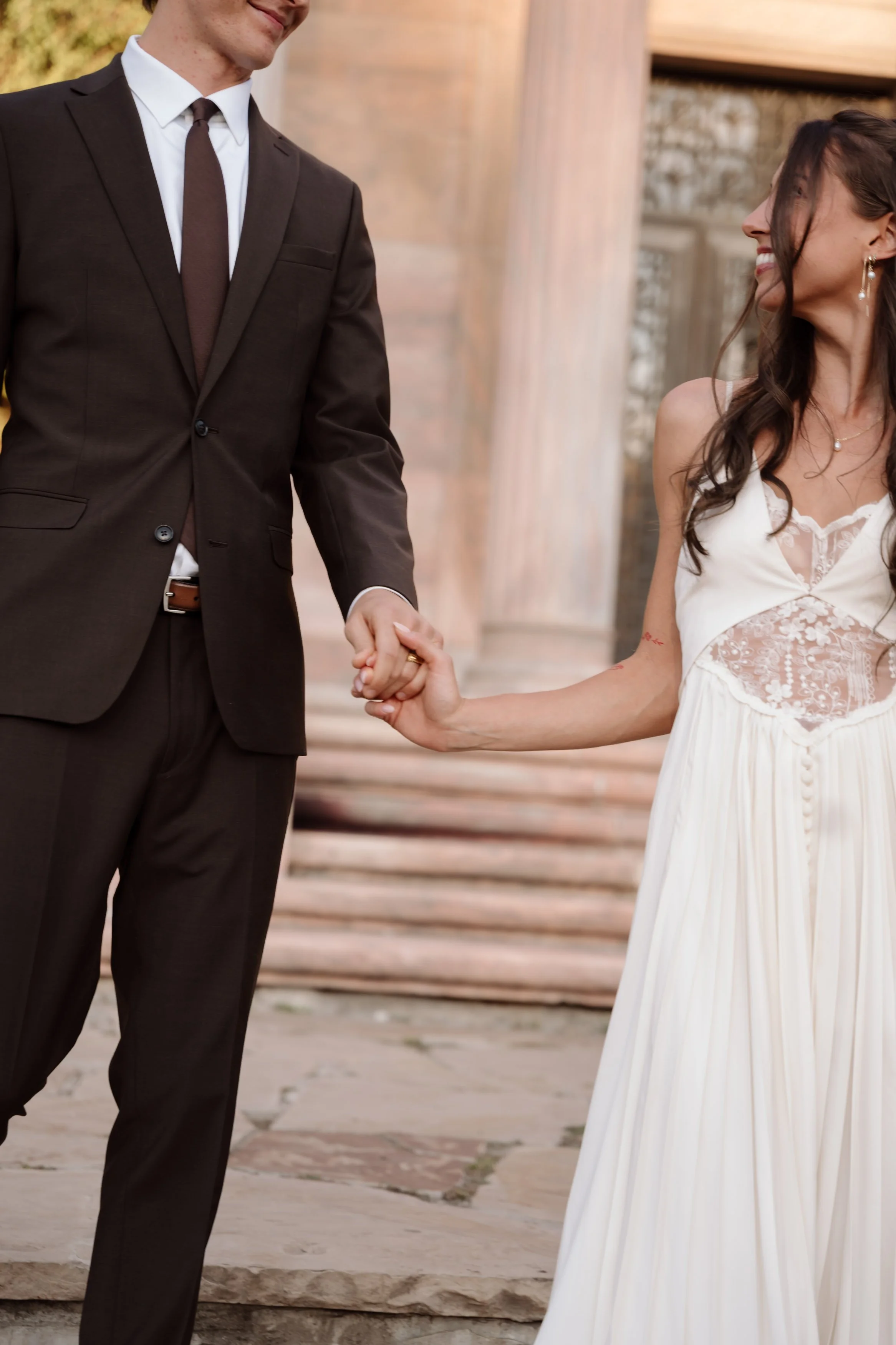 A couple holding hands, with the woman dressed in a white lace and chiffon dress and the man in a dark brown suit, outdoors in front of stone steps and columns.
