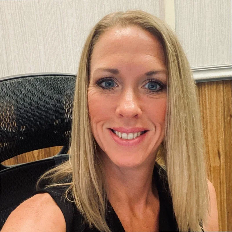 Close-up of a smiling woman with blonde hair and blue eyes in an office setting.